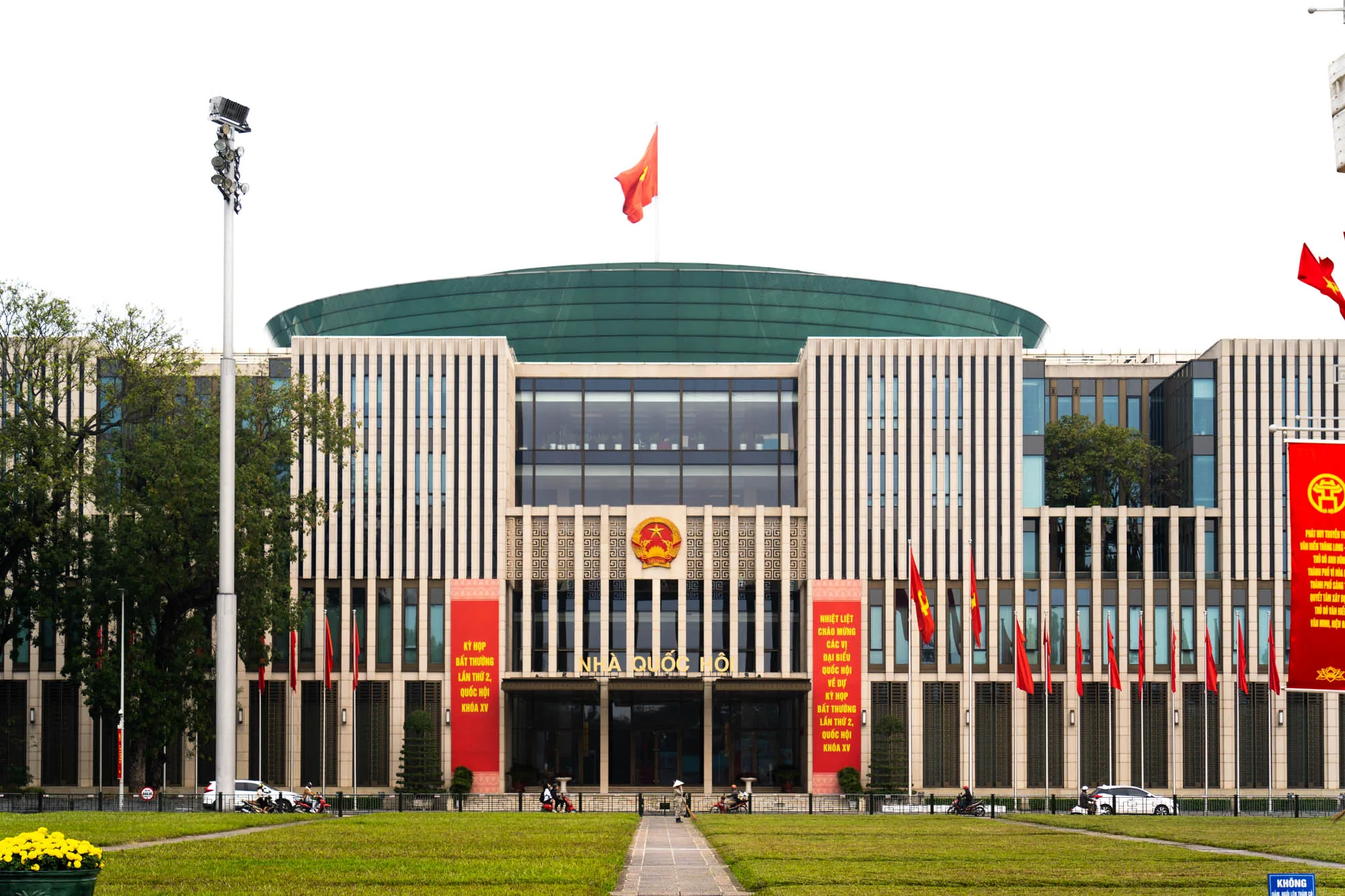 この写真はベトナムのハノイで撮影された国会議事堂の正面を捉えたものです。建物は縦長の窓が整然と並び、その上部に緑色のドーム型の屋根が特徴的です。屋根の上にはベトナムの国旗が掲げられています。建物の正面には「NHÀ QUỐC HỘ」と書かれた文字が見えます。左右には赤いバナーが掲げられ、ベトナム語で「KỲ HỌP BẤT THƯỜNG LẦN THỨ 2, QUỐC HỘI KHÓA XV」（第2回臨時国会、第15期国会）などの文字が書かれています。建物の手前には緑色の芝生が広がり、通路が整備されています。芝生には黄色い花が植えられた鉢植えが置かれています。周囲には街路樹があり、道路を挟んで車やバイクが通行しています。全体的に、厳粛で公式な雰囲気が感じられます。