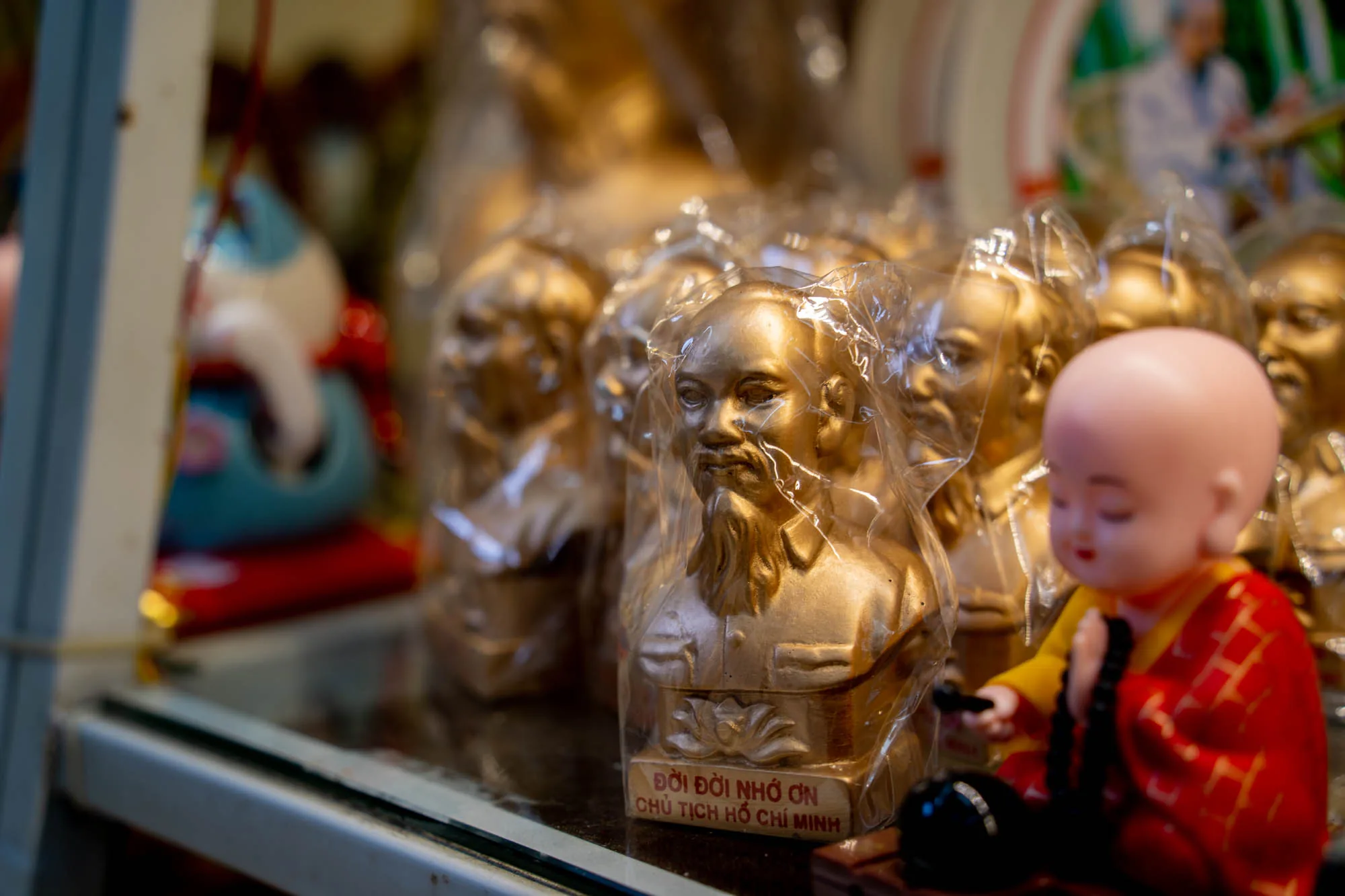 写真には、ベトナムのハノイにある土産物店の一角が写っています。メインの被写体は、黄金色に輝くホー・チ・ミンの胸像が複数並べられた様子です。それぞれの胸像はプラスチックの袋で丁寧に包装されています。胸像の台座には、ベトナム語で「Đời đời nhớ ơn Chủ tịch Hồ Chí Minh」(永遠にホー・チ・ミン主席を偲ぶ)と書かれています。  胸像の隣には、赤い袈裟をまとった小さなお坊さんの人形が座っています。この人形は数珠を手に持ち、穏やかな表情をしています。背景には、ぼやけていますが、他の商品の一部が見えます。全体的に、ハノイの土産物店独特の雰囲気を捉えた一枚です。