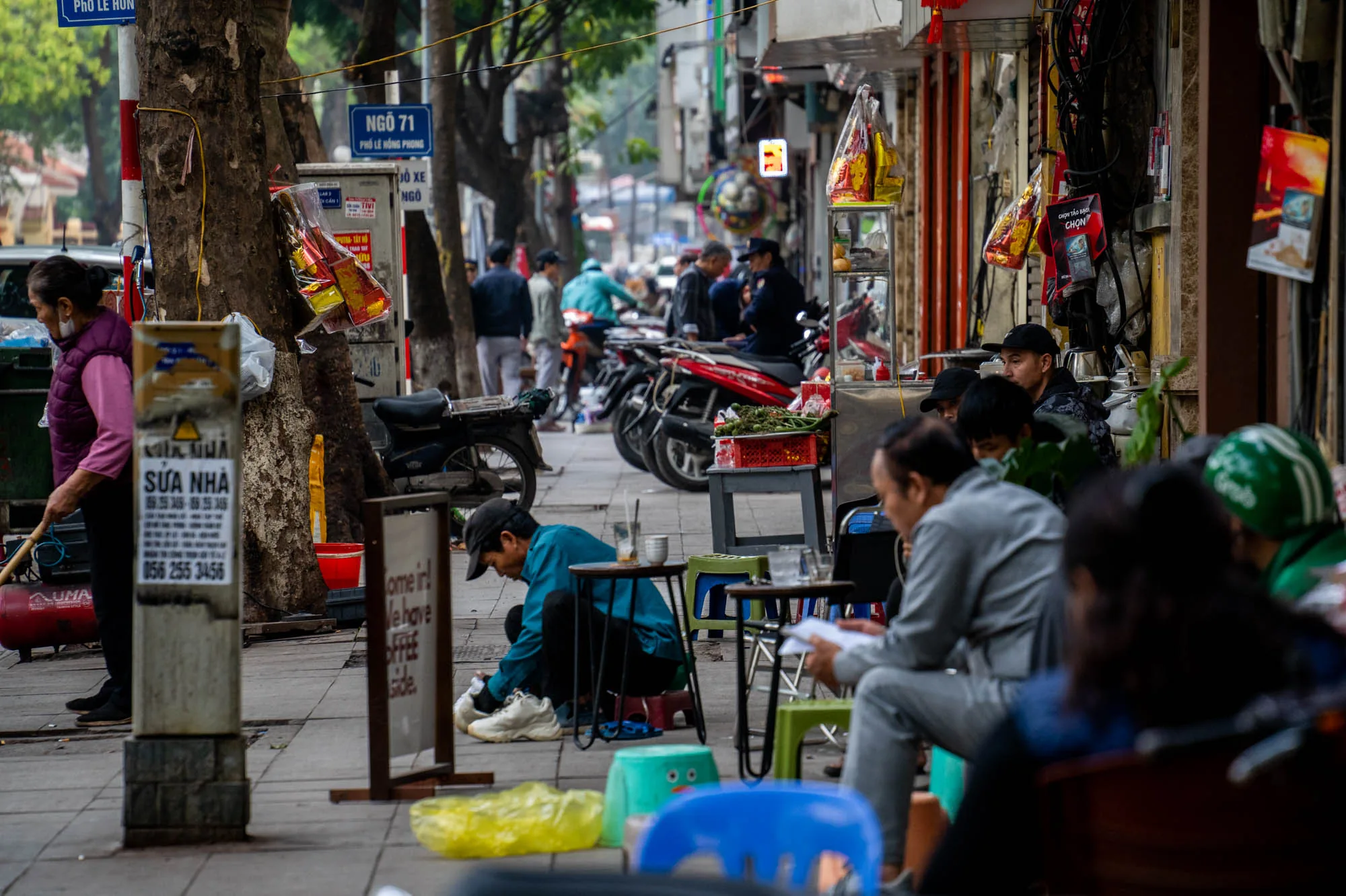 写真はベトナムのハノイで撮影された街並みのスナップショットです。 手前の歩道には、腰を下ろして靴の手入れをしている男性、椅子に座って書類を読んでいる男性、そして、小さな椅子に座って談笑する数人の男性がいます。緑色のヘルメットを被った人物もおり、おそらくバイクタクシーの運転手でしょう。歩道の脇には小さなテーブルとカラフルなプラスチック製の椅子が置かれ、簡易的な飲食スペースになっているようです。  左側には、ほうきを持った女性がいて、近くに赤いコンプレッサーのようなものがあります。電柱には広告や案内が貼られ、その横には木の幹があります。背景にはバイクが数台駐輪されており、人通りも多いことが伺えます。お店の軒先には商品や飾り物が吊るされ、活気のある雰囲気を醸し出しています。 全体的に落ち着いた色調で、ハノイの日常の喧騒と人々の生活感が伝わってくる写真です。