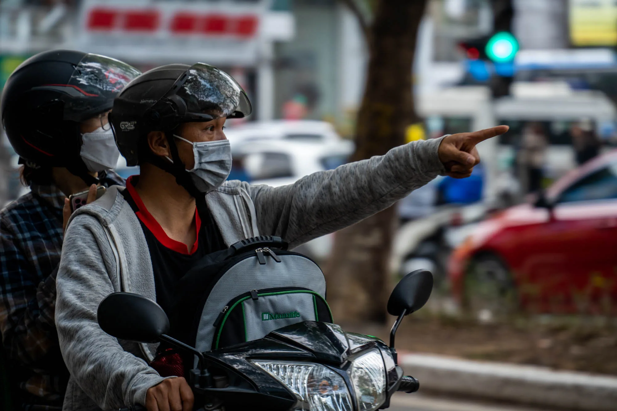 ベトナムのハノイで撮影された写真です。バイクに乗った二人の人物が写っています。運転手はマスクとヘルメットを着用しており、助手席の人物も同様の格好をしています。運転手は右手で何かを指し示しており、道案内をしている様子が伺えます。バイクの前には「Manulife」と書かれたバッグが取り付けられています。背景には、信号機が緑色に点灯しているのが見え、他の車両や建物がぼんやりと写っています。写真全体からは、ハノイの日常的な交通風景が伝わってきます。