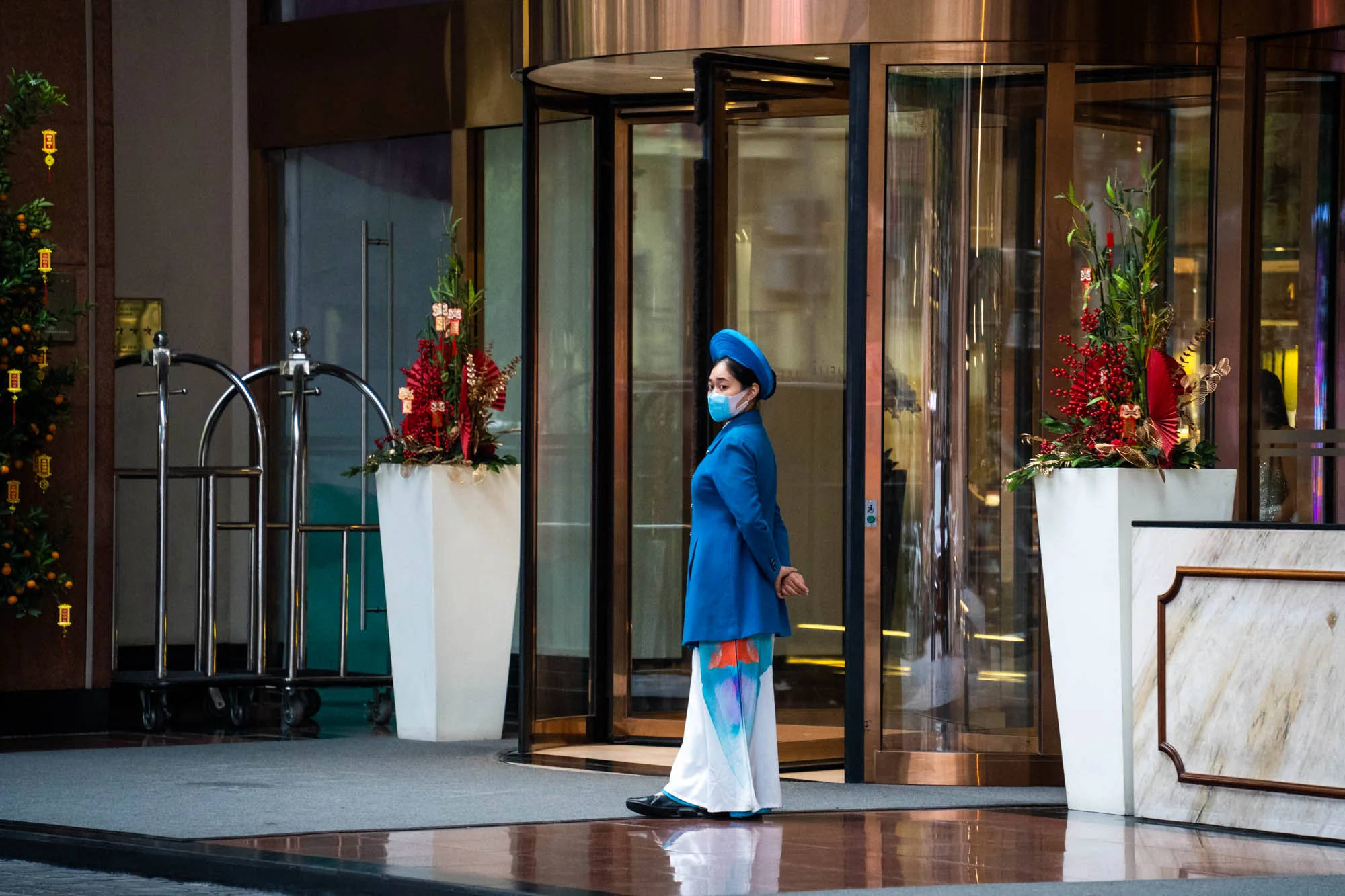 ベトナムのハノイで撮影された写真です。ホテルの入り口付近が写っており、回転ドアの手前に、伝統的な衣装であるアオザイを着た女性が立っています。女性は青色のジャケットと帽子を身につけ、マスクを着用しています。彼女は両手を後ろに組み、静かに立っている様子です。背景には、ホテル内部の様子がわずかに見え、ロビーには荷物運搬用のカートや、正月飾りと思われる花などが飾られています。床は磨かれており、光沢があり、反射しています。全体的に、高級ホテルで働く従業員が、職務を遂行している場面を捉えた写真と言えるでしょう。