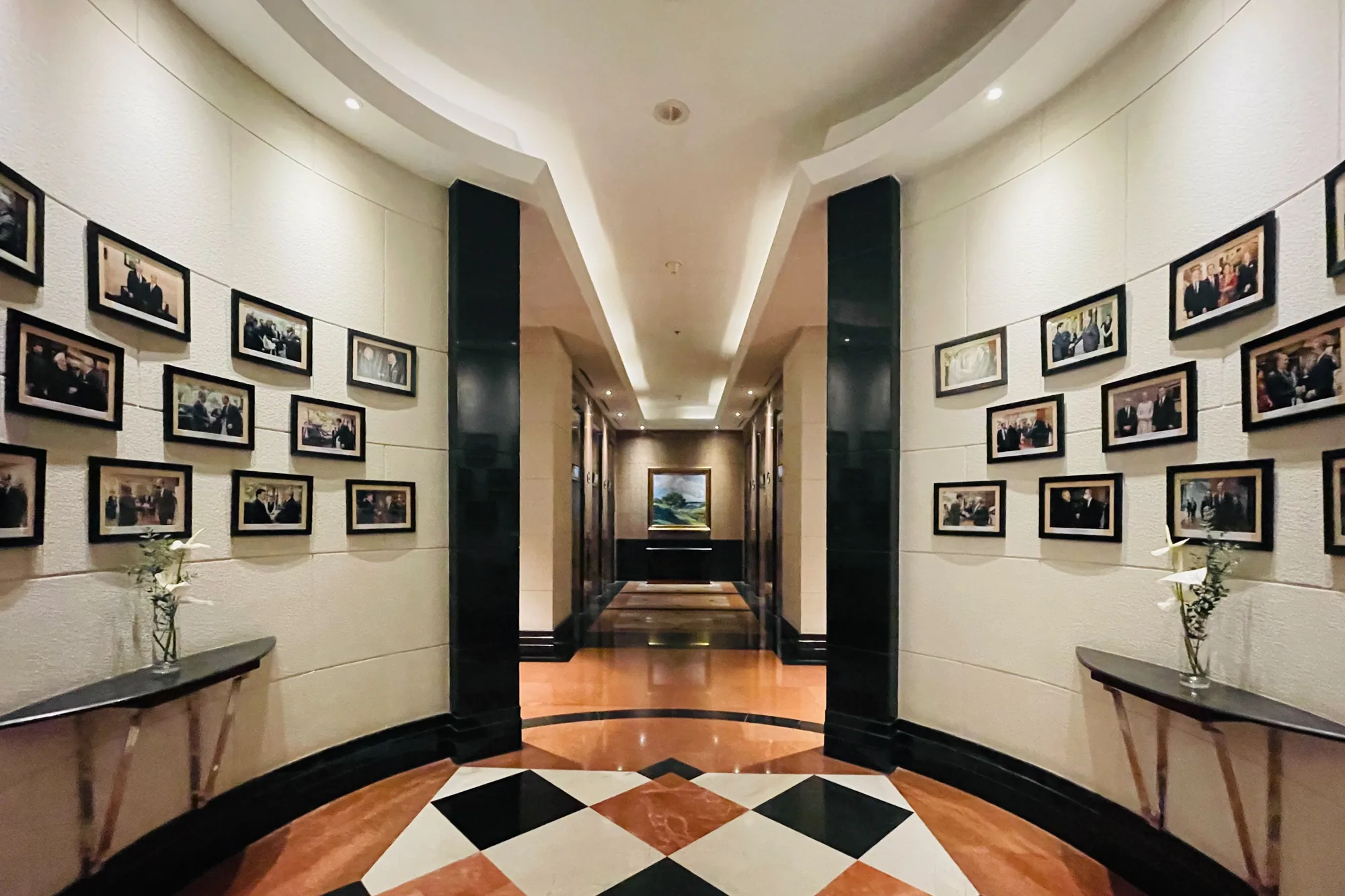 ベトナムのハノイで撮影された、豪華な内装の廊下の写真です。廊下の入り口部分は緩やかな曲線を描いており、壁には黒いフレームに入れられた人物の写真が複数飾られています。写真の下には、花瓶に生けられた白い花が飾られたコンソールテーブルが置かれています。廊下の奥へ進むにつれて直線的なデザインに変わり、床は茶色と白の菱形模様のタイル張りになっています。廊下の突き当たりには絵画が飾られており、その下にはテーブルが置かれています。照明は天井に埋め込まれたライトによって照らされ、落ち着いた雰囲気を作り出しています。全体的に、高級ホテルや格式高い施設の内部を思わせる、洗練された空間を捉えた一枚です。