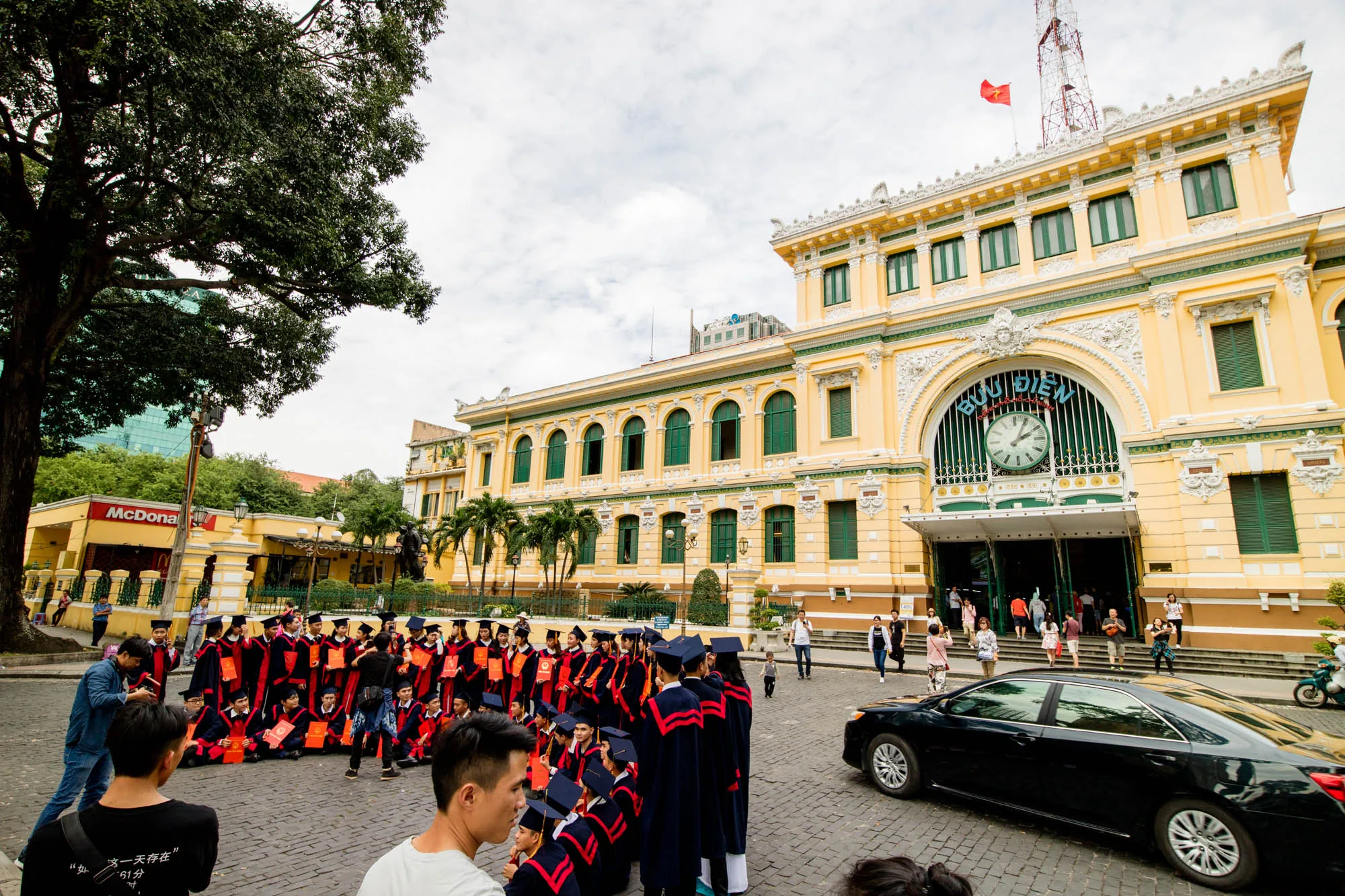 ベトナムのホーチミン市中央郵便局前で、卒業式を迎えた学生たちが記念撮影をしている風景です。学生たちは、黒いガウンに赤いラインが入った卒業衣装を身につけ、卒業証書らしき赤い筒状のものを手にしています。一部の学生は座り、一部は立って、カメラに向かってポーズをとっています。背景には、黄色い外壁が特徴的なホーチミン市中央郵便局が写っており、その上にはベトナムの国旗が掲げられています。郵便局の前には人が行き交い、路上には黒い車が停車しています。左手にはマクドナルドの看板も見え、周辺が賑わっている様子が伺えます。画面左側には大きな木があり、緑豊かな雰囲気を添えています。全体的に、卒業の喜びと未来への希望に満ちた、明るい雰囲気の写真です。