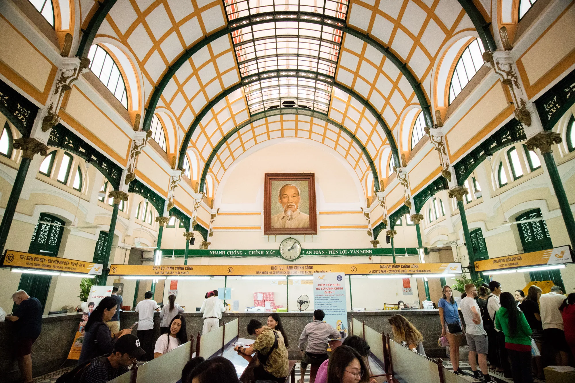 写真はベトナムのハノイにある郵便局の内部を捉えています。建物は高く、アーチ状の天井が印象的です。天井は格子状になっており、自然光が差し込んでいます。壁はクリーム色で、装飾的な緑色の柱が並んでいます。正面にはホー・チ・ミン主席の肖像画が飾られています。  窓口には多くの人が並んでおり、現地の人が様々な手続きを行っている様子が伺えます。窓口には番号が振られており、それぞれ異なるサービスを提供しているようです。写真全体は活気があり、歴史的な建物の中で人々が日常的な活動を行っている様子が伝わってきます。建物自体の装飾や構造、人々の服装から、ハノイという土地の文化と歴史を感じられる一枚です。
