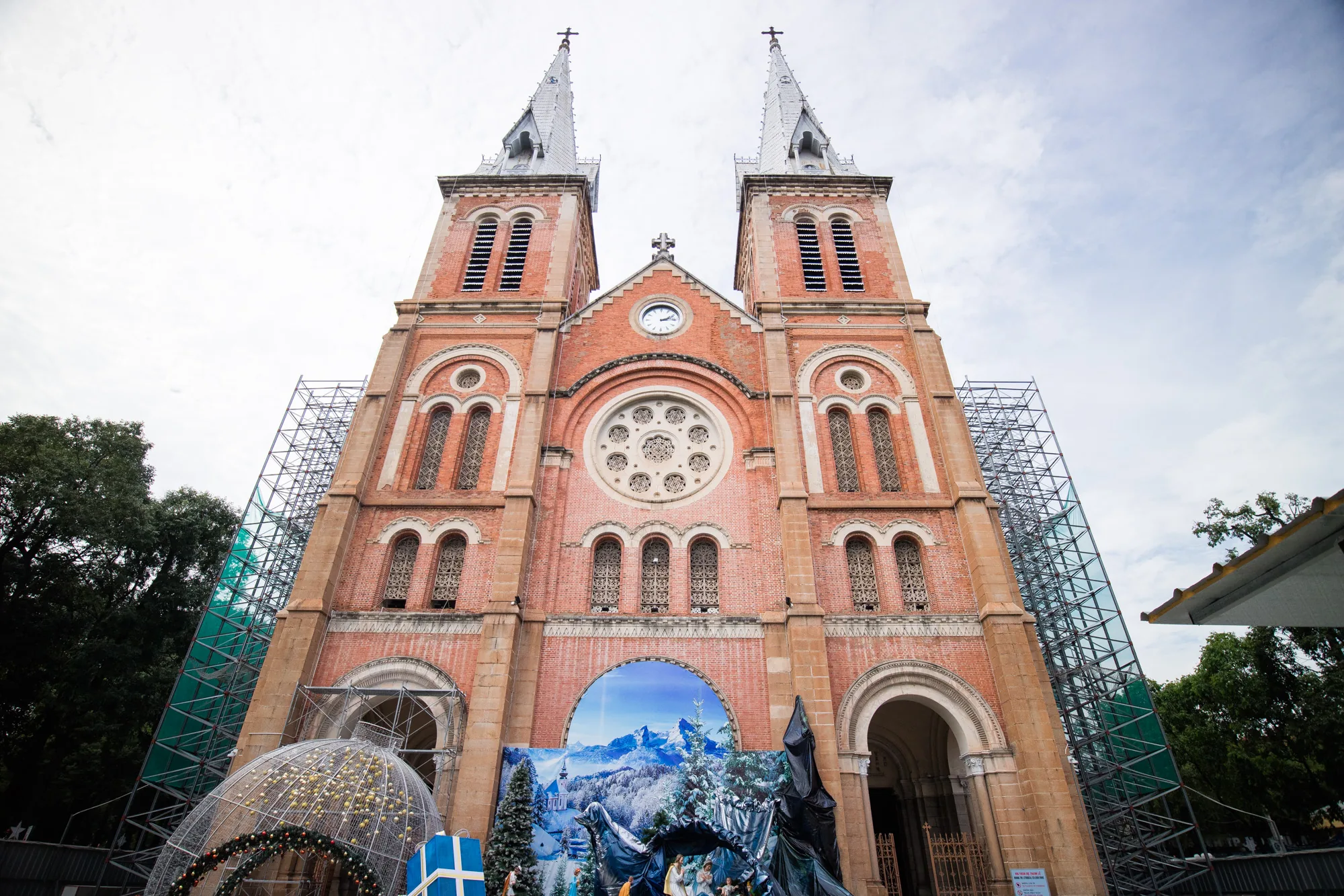 ベトナムのハノイにある、レンガ造りの荘厳な教会の写真です。教会正面には2つの高い尖塔がそびえ立ち、中央には時計が設置されています。建物の外観は補修工事中なのか、一部に足場が組まれています。教会の前には、冬の風景が描かれた大きなバナーが飾られ、クリスマスツリーや馬小屋のジオラマが設置されています。また、大きなオーナメントボール型のイルミネーションやプレゼントボックスも置かれており、クリスマスシーズンであることがわかります。空は曇り空で、落ち着いた雰囲気の写真です。