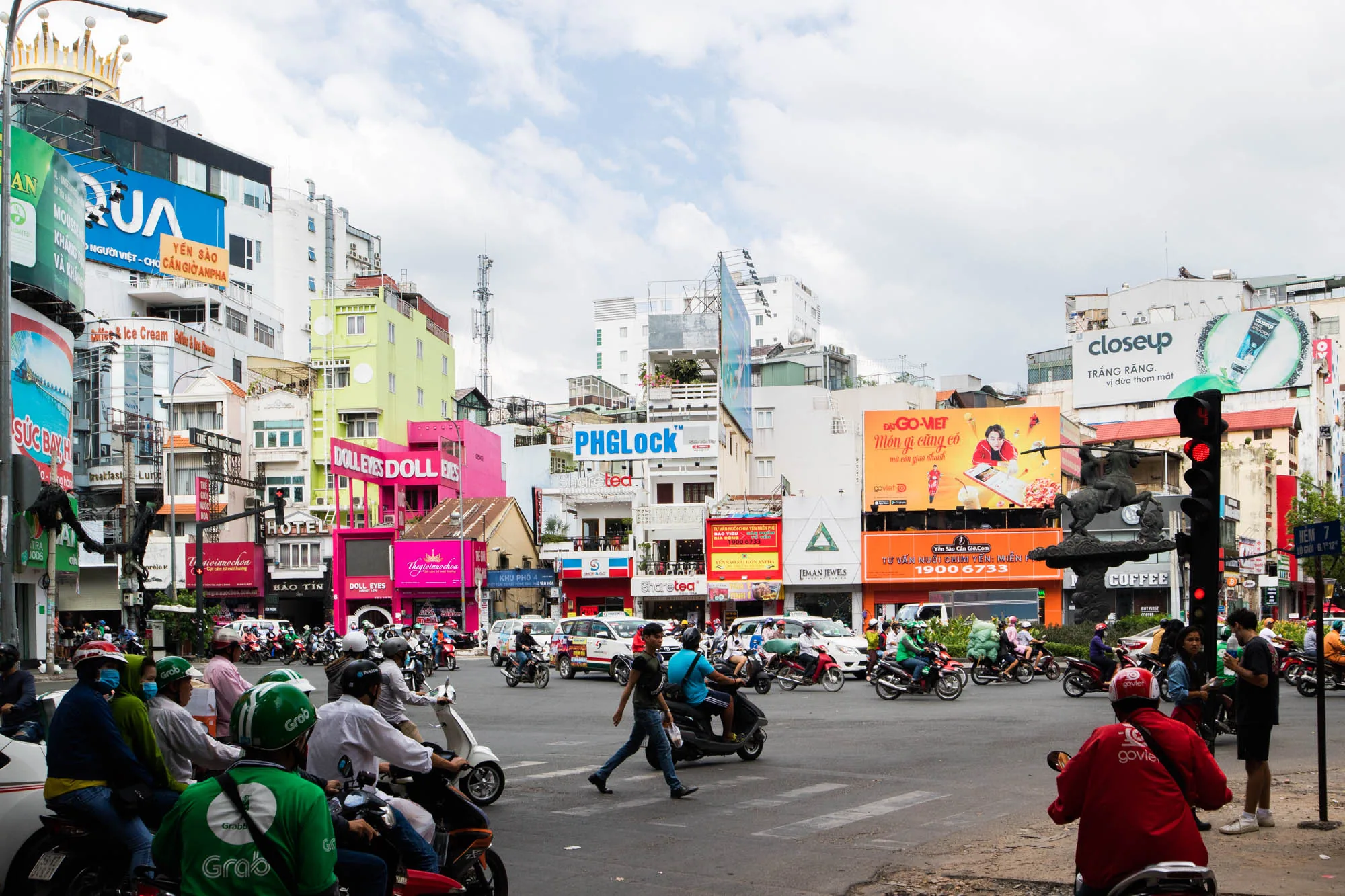 写真はベトナムのハノイで撮影された街の風景です。交差点に多くのバイクや車が走っており、活気に満ちた様子が伝わってきます。Grabの配達員と思われる緑色の制服を着た人々が複数写っており、背景には様々な色の建物や広告看板が密集しています。ピンクや黄緑色の建物が目を引き、UAやcloseupといったブランドの広告看板が見られます。また、道の反対側にはホテルと思われる建物や、コーヒーショップの看板も確認できます。空は薄曇りで、全体的に少し霞んだような雰囲気です。信号が赤なので、バイクが停止しており、歩行者が横断歩道を渡っています。交通量が多く、騒がしい街の雰囲気が伝わってきます。