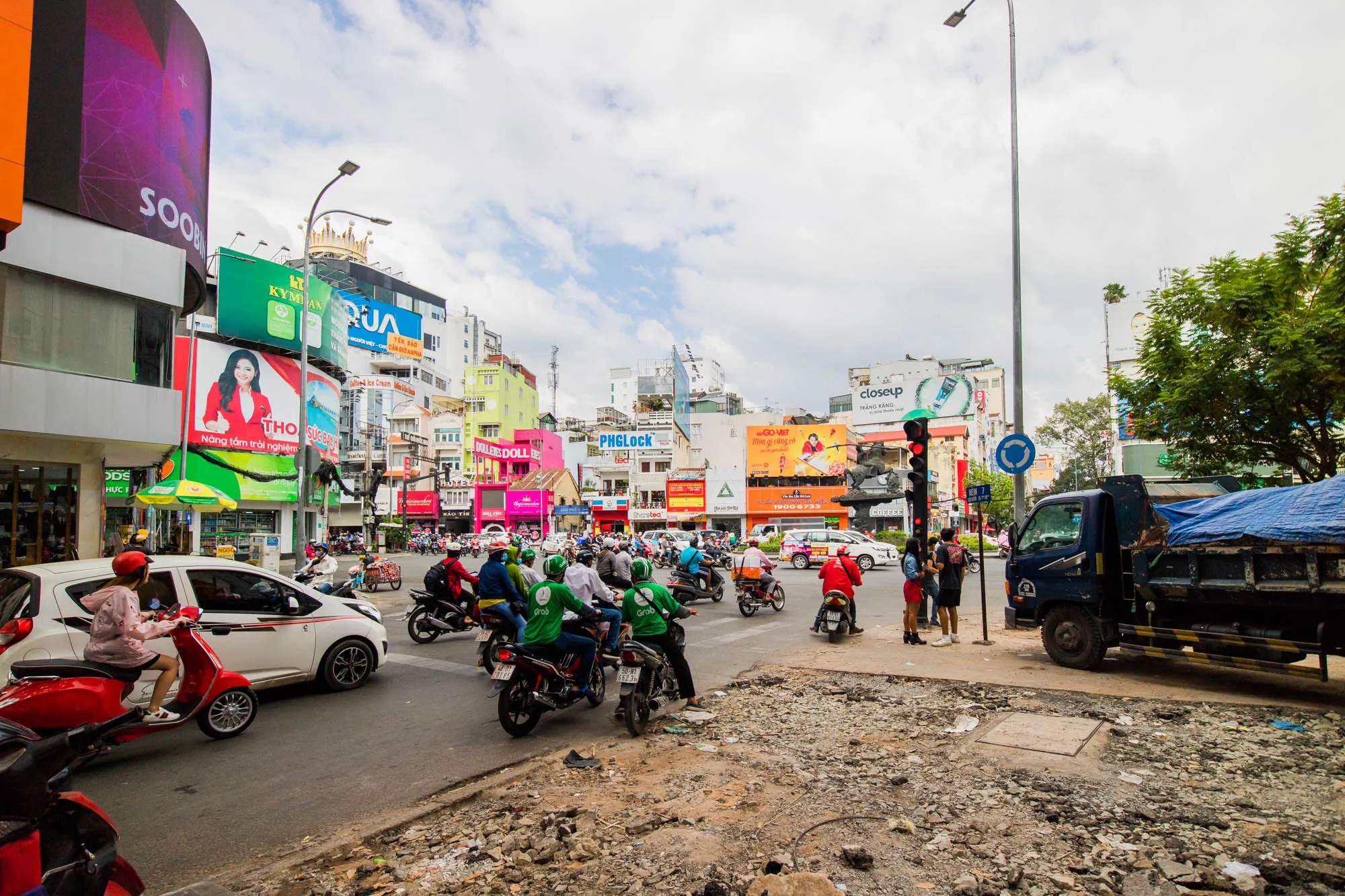 写真はベトナムのハノイの賑やかな交差点を捉えています。多くのバイクや車が行き交い、活気に満ちています。特にGrabの緑色のユニフォームを着たバイクタクシーの運転手たちが目立ちます。背景には様々な広告看板が立ち並び、カラフルな建物が密集しています。空は曇り空で、路上は多少荒れているように見えます。歩道には人々が立っており、トラックが停車している様子も写っています。全体的に、ハノイの日常の喧騒と活気を伝えている写真です。