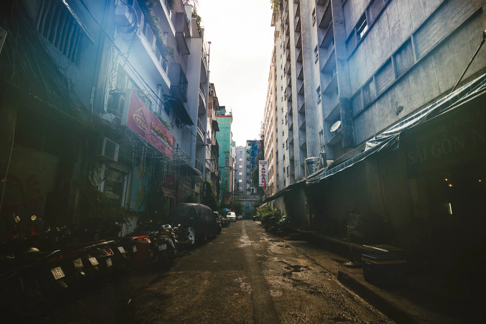 写真はベトナムのハノイで撮影された路地裏の風景です。狭い路地は、左右を高い建物に挟まれており、空は白く霞んでいます。左側の建物は比較的低層で、エアコンの室外機や看板、植物が密集しており、生活感があふれています。看板には「KHACH SAN」、「SÀI GÒN」などの文字が見えます。路地の左側には、多くのバイクが縦列駐車されています。右側の建物は高層で、コンクリート打ちっぱなしのような外観です。路面は濡れており、ところどころに水たまりがあります。全体的に暗く、レトロな雰囲気が漂っています。路地の奥には白い車が見え、人々の生活の匂いが感じられます。