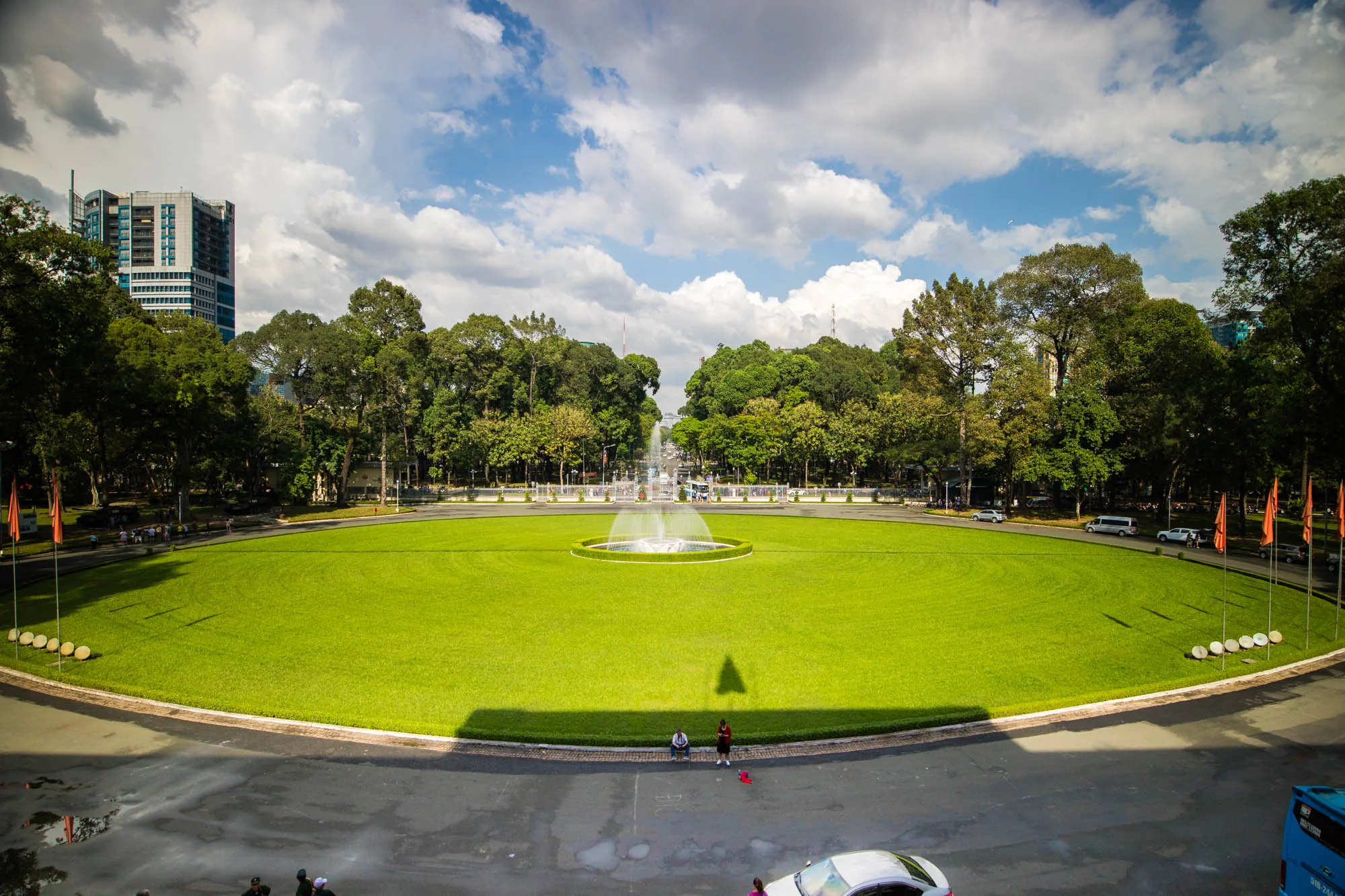 写真には、ベトナムのハノイで撮影されたと思われる、広大で手入れの行き届いた公園が写っています。画面中央には、明るい緑色の芝生が広がり、その中心には水しぶきを上げる噴水があります。公園の奥には木々が生い茂り、さらに遠くには高層ビルがそびえ立っています。空は青く、白い雲が点在しており、明るい日差しが公園全体を照らしています。前景には舗装された道路があり、数人の人が座ってくつろいでいたり、車が停まっていたりします。全体的に、穏やかで開放感のある雰囲気の写真です。
