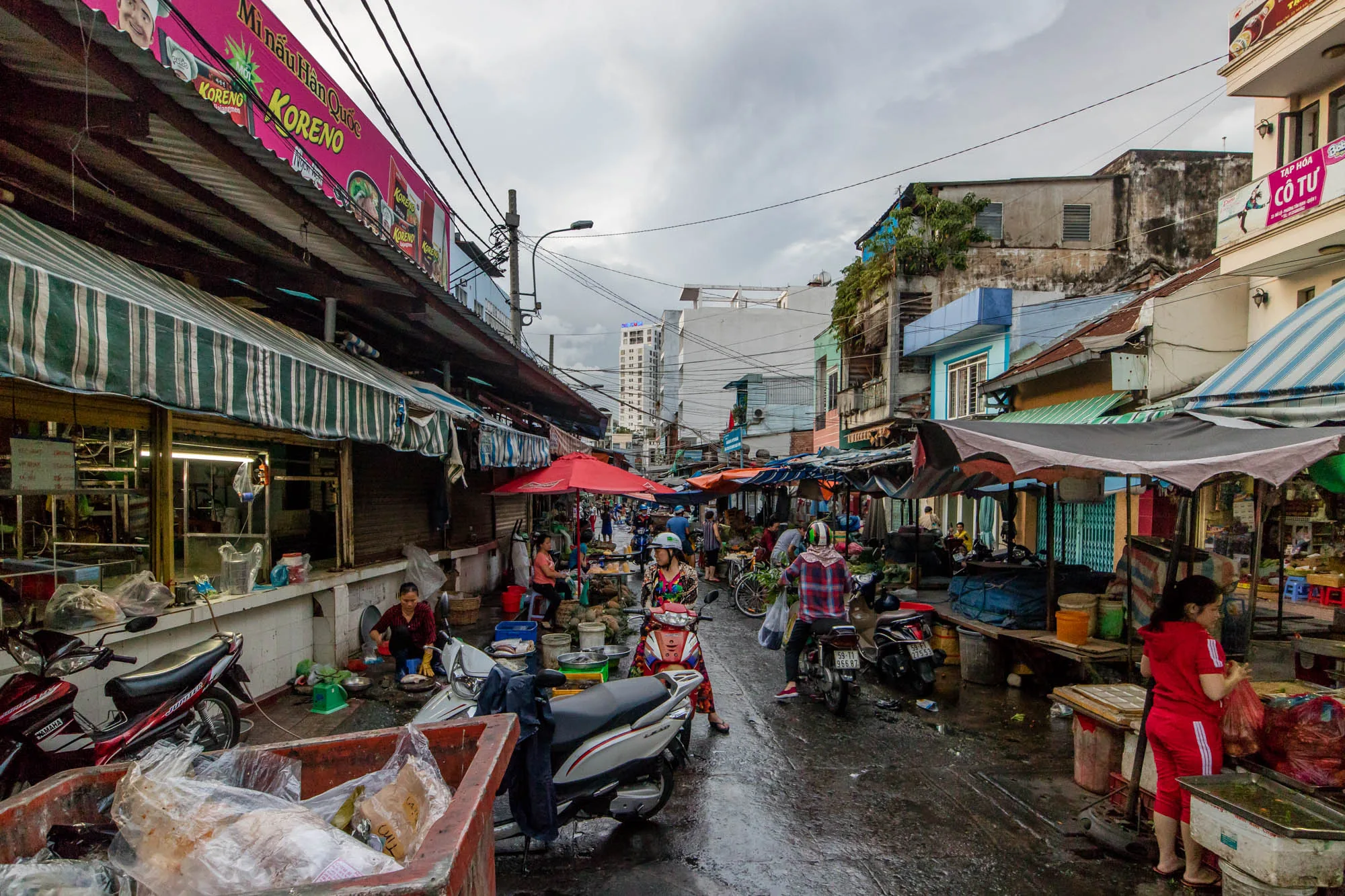 写真はベトナムのハノイで撮影された市場の様子を捉えています。雨上がりの濡れた路面が特徴的で、曇り空の下、活気のある市場の雰囲気が伝わってきます。道路の両脇には、屋根や庇の下に様々な店が並び、人々が買い物や商売をしています。バイクが複数台駐車されており、人々の移動手段として利用されている様子がうかがえます。看板にはベトナム語の文字が見られ、ローカルな雰囲気を醸し出しています。奥には高層ビルが見え、都市部の市場であることが分かります。全体的に混沌としていますが、それがまた独特の魅力を放っている一枚です。鮮やかな赤色の傘や衣服が、写真全体の彩りを添えています。