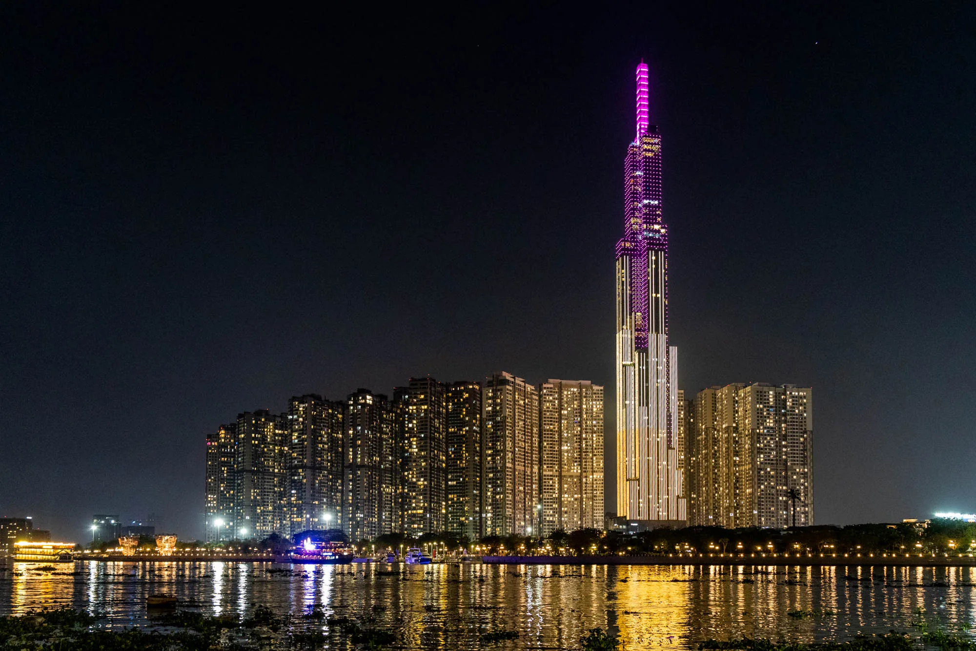 ベトナムのホーチミン市で撮影された夜景の写真です。画面奥には、ピンク色にライトアップされた非常に高い高層ビルがそびえ立っています。このビルはランドマーク的な存在で、多くの人々の目を引くことでしょう。その手前には、複数の高層マンションが立ち並び、各戸の窓から漏れる光が、水面に反射して輝いています。画面手前には川が写っており、水面は穏やかで、光が揺らめいています。川沿いには遊歩道のようなものが見え、街灯が並んでいます。雰囲気は全体的に落ち着いており、都会の夜の静けさを感じさせます。