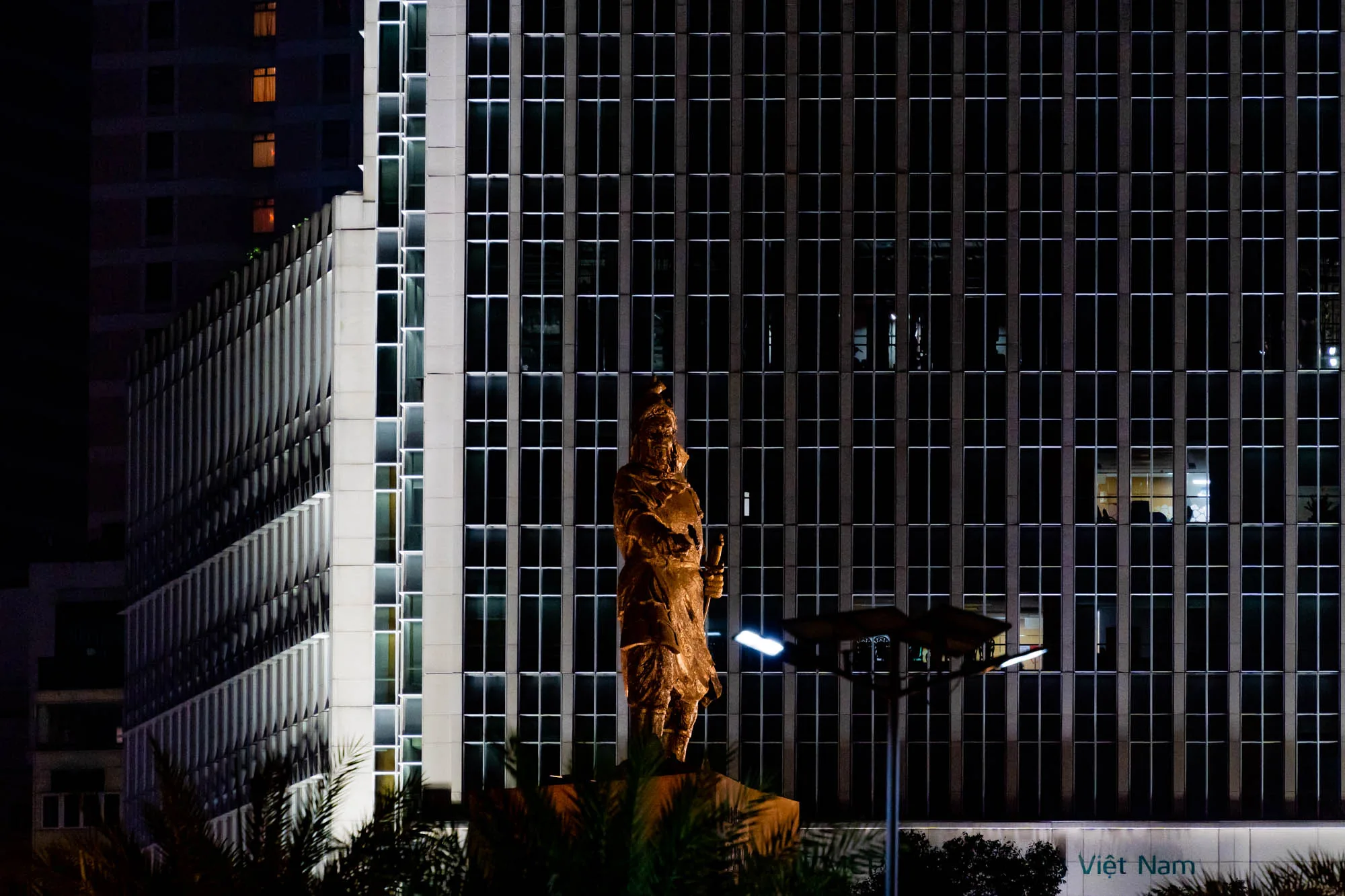 写真はベトナムのホーチミンで撮影された夜景です。中央には、古代の服装を身につけた人物の像が立っており、ライトアップによって金色に輝いています。像は、近代的な高層ビルの前に位置しており、建物の壁面は規則的な窓の配列で構成されています。窓のいくつかには明かりが灯っており、人影も見えます。手前の植物はシルエットとして浮かび上がり、夜の雰囲気を醸し出しています。全体的に、歴史的な要素と現代的な都市景観が組み合わさった、コントラストの強い写真です。
