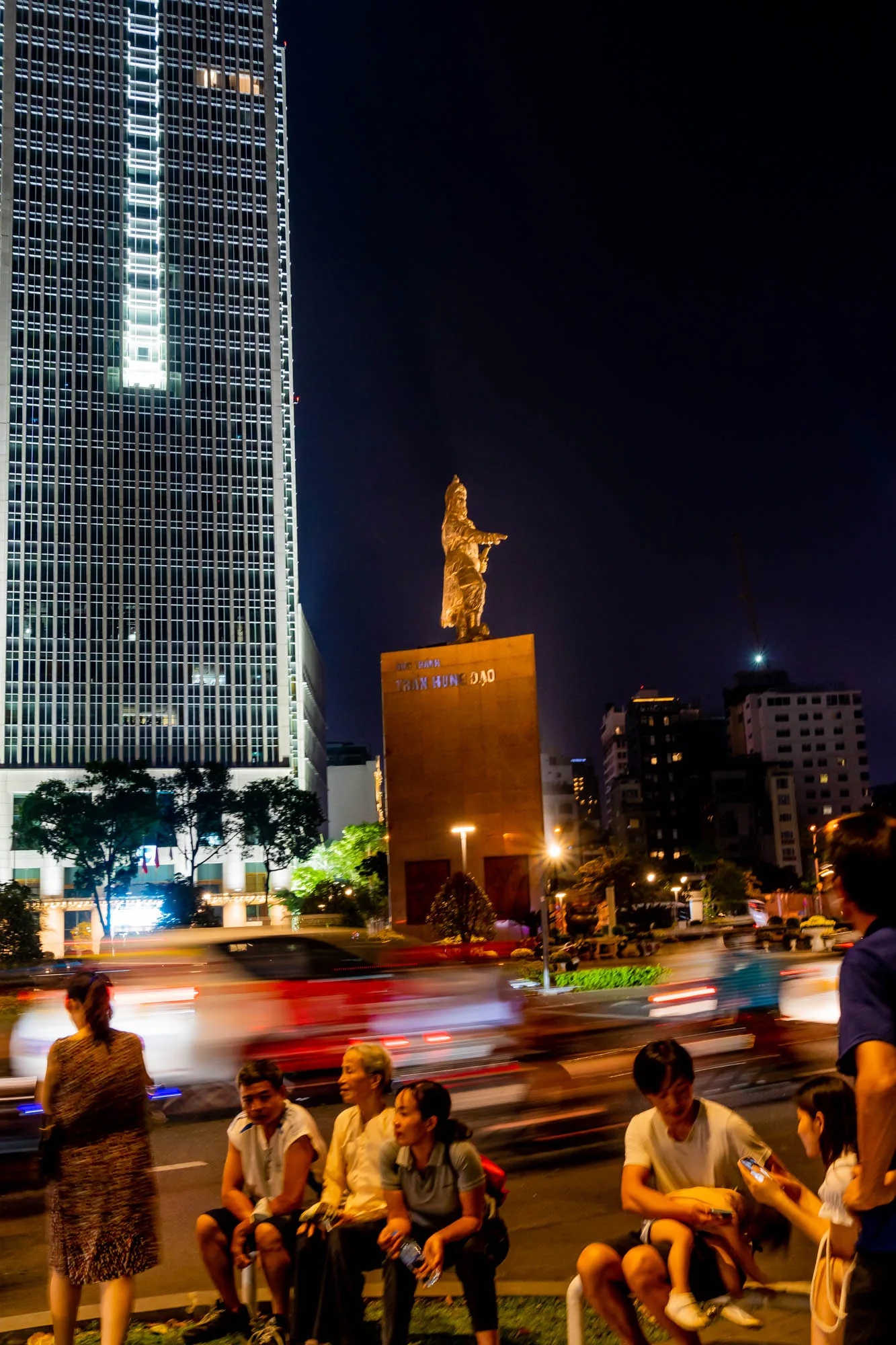 この写真はベトナムのホーチミン市で撮影された夜景です。画面左側には高層ビルがそびえ立ち、ライトアップされています。中央にはチャン・フン・ダオの像がライトアップされ、威厳を放っています。像の前には人々が座って休憩しており、その前を車両のライトが光の軌跡を描きながら通り過ぎています。夜の街の賑わいと、人々の穏やかな時間が組み合わさった様子が捉えられています。
