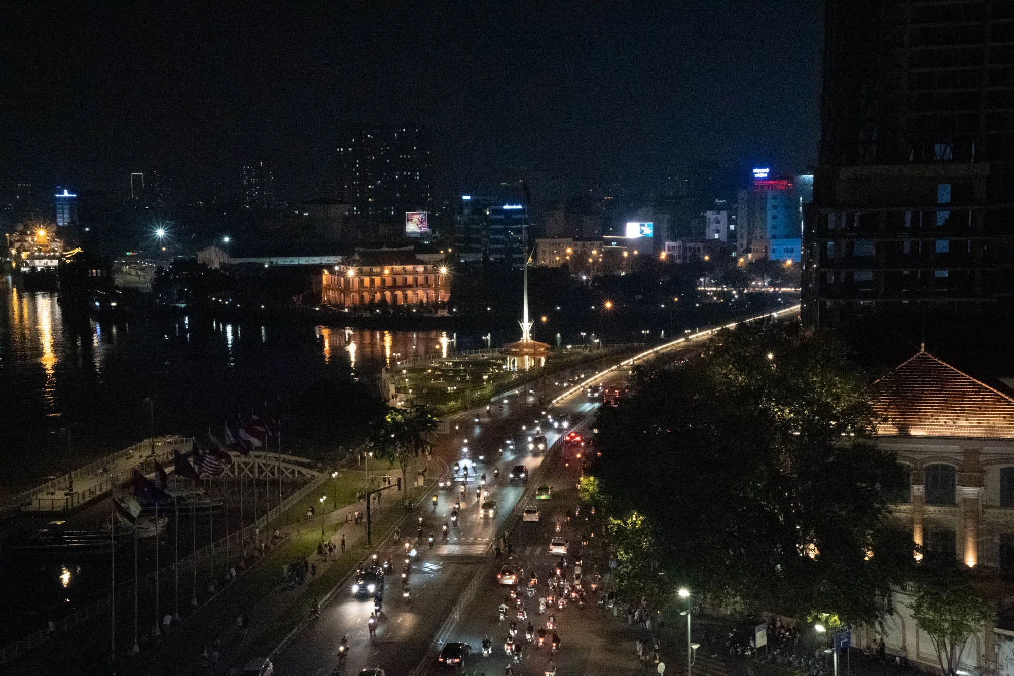 ベトナムのホーチミン市で撮影された夜景の写真です。暗闇の中に、川沿いの建物や道路がライトアップされ、美しい光景が広がっています。川には船が停泊しており、その明かりが水面に反射しています。道路には無数のバイクや車のヘッドライトが連なり、活気ある街の様子を伝えています。遠くには高層ビル群の明かりも見え、ホーチミン市の発展を象徴しているようです。手前には大きな木々があり、夜の静けさを感じさせます。全体的にノイズが多く暗い写真ですが、夜のホーチミン市の魅力を捉えた一枚と言えるでしょう。