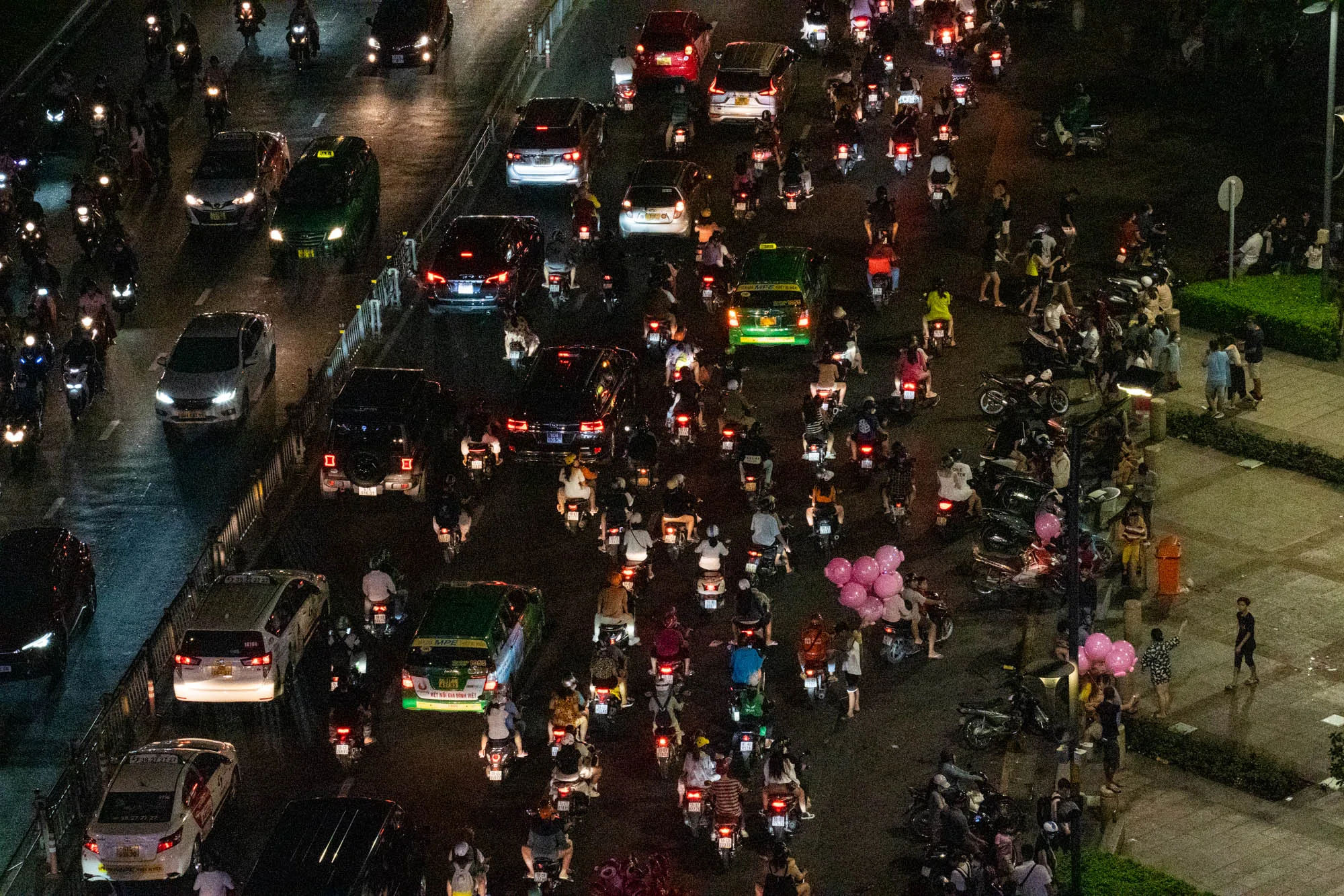 この写真はベトナムのホーチミン市で撮影された夜景です。高い場所から見下ろした構図で、道路には大量のバイクと車がひしめき合っています。車やバイクのヘッドライトとテールランプが輝き、交通量の多さを物語っています。道路脇の歩道には人が集まっており、一部の人はピンクの風船を持っています。夜の賑やかな雰囲気と、ベトナム特有の交通事情が伝わってくる一枚です。OCRで読める範囲では、タクシー会社の情報や「KẾT NỐI GIA ĐÌNH VIỆT（ベトナムの家族のつながり）」という言葉も確認できます。