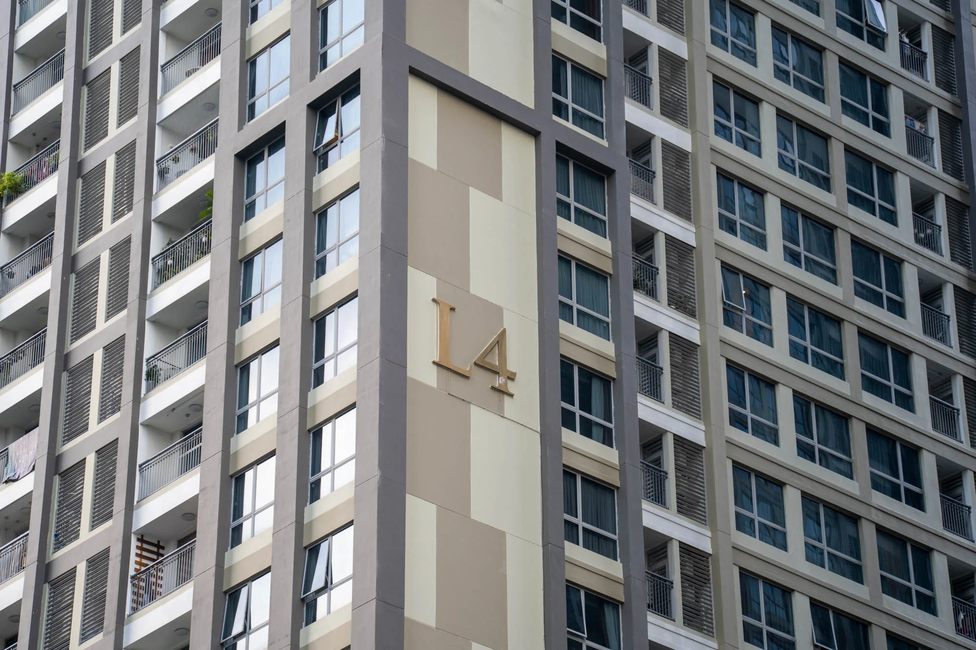 写真は、ベトナムのホーチミン市で撮影された高層マンションの一部を捉えています。建物の外観は、グレーを基調としたモダンなデザインで、規則正しく配置された窓とバルコニーが特徴的です。バルコニーには植物が置かれているところもあり、生活感を感じさせます。マンションの角の部分には、「L4」という文字が大きく表示されています。全体的に落ち着いた色調で、都市の景観を構成する一部として、静かに佇んでいる様子が伝わってきます。建物の窓には周囲の景色が反射しており、周囲の建物や空の様子が映り込んでいます。
