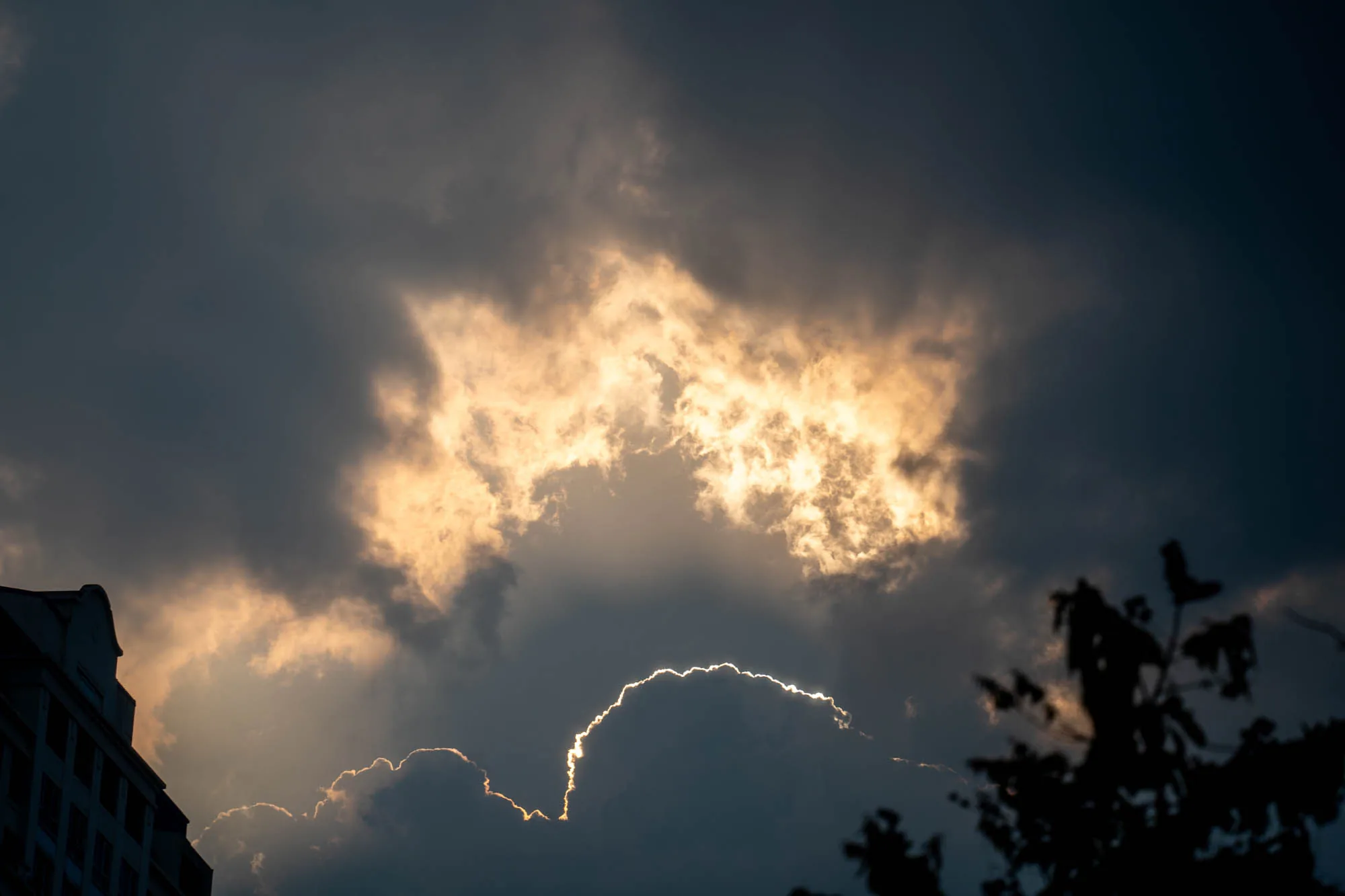 この写真は、ベトナムのホーチミンで撮影された、劇的な空模様を捉えたものです。画面全体を覆うように、厚く重い雲が広がっています。雲の色は暗いグレーから黒に近い色まであり、全体的に陰鬱な雰囲気を醸し出しています。しかし、その雲の隙間から、強い太陽光が差し込んでおり、雲の輪郭を金色に縁取っています。特に、画面中央付近の雲は、その光によって内側から照らされているかのように明るく輝いています。画面左端には、建物の輪郭の一部が見えます。また、画面右端には、木々のシルエットが写っています。全体的に、光と影のコントラストが印象的で、神秘的な雰囲気を感じさせる写真です。