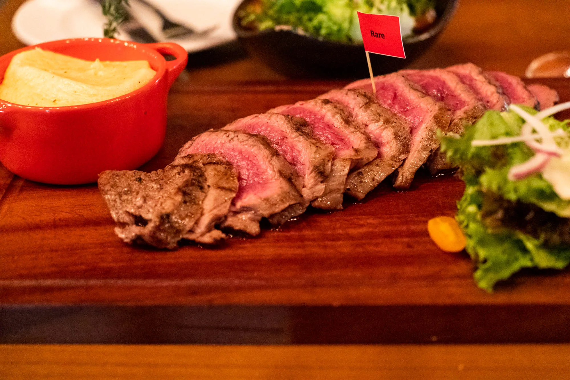 写真には、木製のまな板の上に盛り付けられたステーキディナーが写っています。ステーキは薄くスライスされており、内部は鮮やかなピンク色で、レアで調理されていることを示しています。ステーキには「Rare」と書かれた小さな赤い旗が刺さっています。ステーキの隣には、小さな赤い容器に入ったマッシュポテト、サラダが添えられています。背景には、別のサラダボウルとぼやけた食器が見えます。全体的に、食事は食欲をそそるように提示されており、高級レストランの雰囲気を感じさせます。