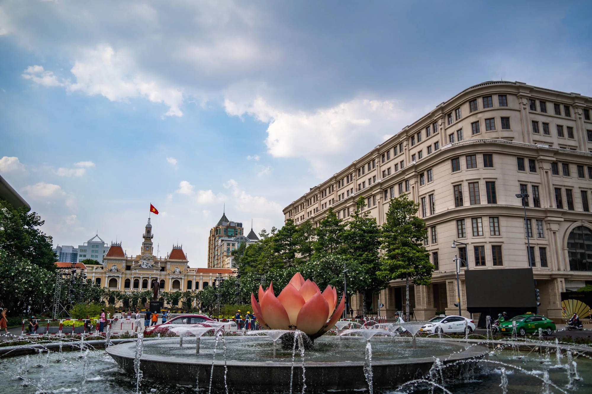写真はベトナムのホーチミン市で撮影されたもので、青空と白い雲が広がる穏やかな日の様子が写っています。前景には噴水があり、中央に蓮の花のオブジェが設置されています。噴水からは水が勢いよく噴き出し、涼しげな雰囲気です。背景には歴史的な建物や高層ビルが並び、ホーチミン市の景観を形成しています。左奥にはベトナムの国旗が掲げられた建物が見え、その手前には人々が歩いています。右側にはベージュ色の建物がそびえ立ち、現代的な雰囲気を醸し出しています。道路にはタクシーやバイクが走り、活気のある街の様子が伝わってきます。全体的に、ホーチミン市の歴史と現代が融合した風景が美しく表現されています。