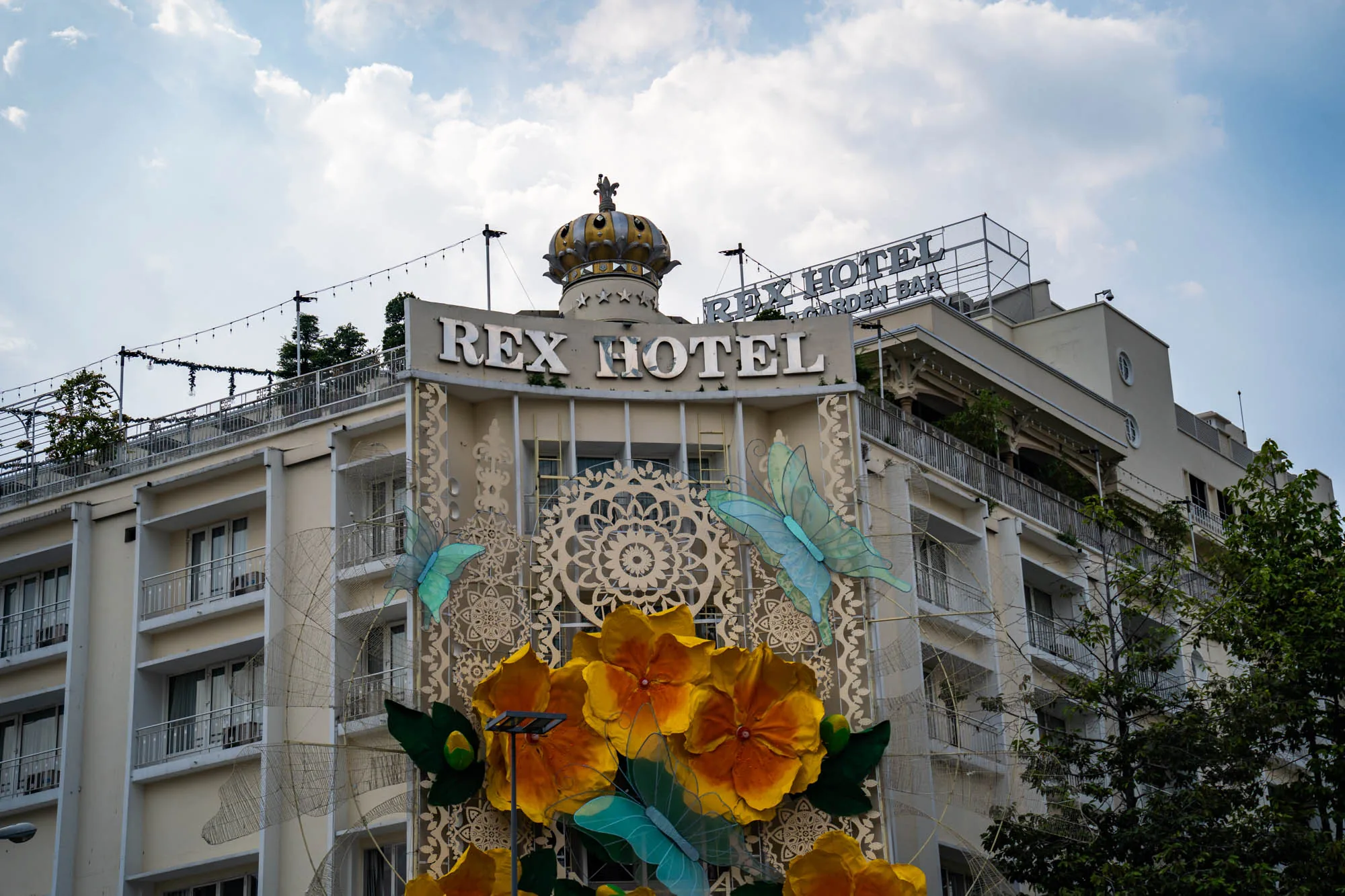 ホーチミンにあるレックスホテルの外観を捉えた写真です。ホテルは白を基調としたクラシックなデザインで、複数の階層があります。屋上には庭園のようなスペースがあり、植物が見えます。ホテルの正面には「REX HOTEL」という大きな文字が掲げられており、その上には王冠の装飾が施されています。ホテルの壁面には、大きな花や蝶の装飾が取り付けられており、華やかな雰囲気を醸し出しています。空は薄曇りで、全体的に落ち着いた色調の写真です。ホテルの一部は木々に隠れており、街中のホテルであることがうかがえます。