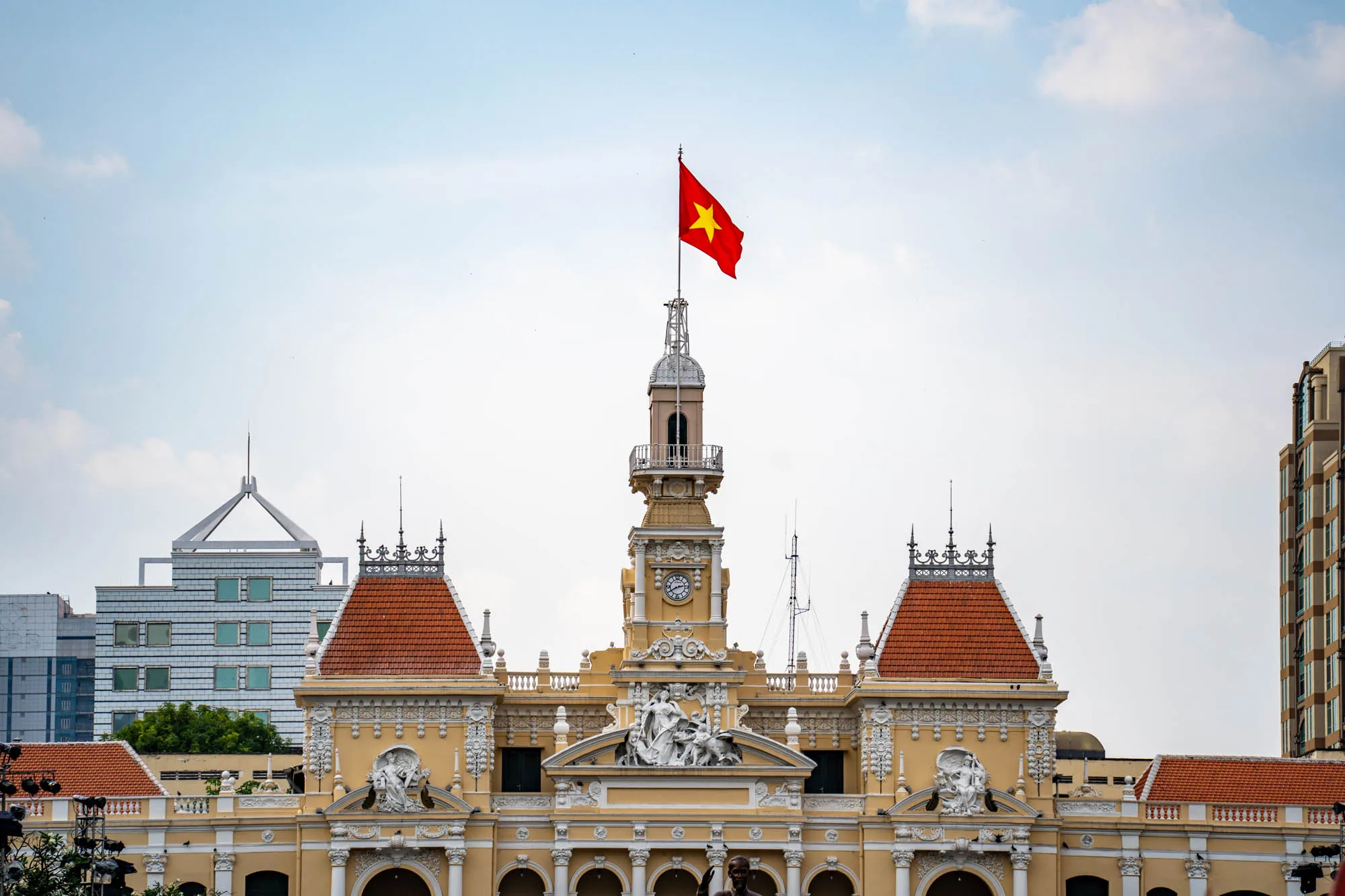 写真は、ベトナムのホーチミン市にある人民委員会の庁舎（旧サイゴン市庁舎）を捉えています。クリーム色の外観で、オレンジ色の屋根瓦が特徴的な美しい建物です。建物の上部には時計塔があり、その頂上にはベトナム国旗が掲げられています。空は薄い水色で、建物と旗が鮮やかに映えています。背景には近代的なビル群も写っており、歴史的な建造物と現代的な都市景観が組み合わさったコントラストが印象的です。建物の装飾は細部にまで凝っており、彫刻やアーチが施されています。全体的に、ホーチミン市のランドマークとしての存在感を示す、堂々とした雰囲気の写真です。