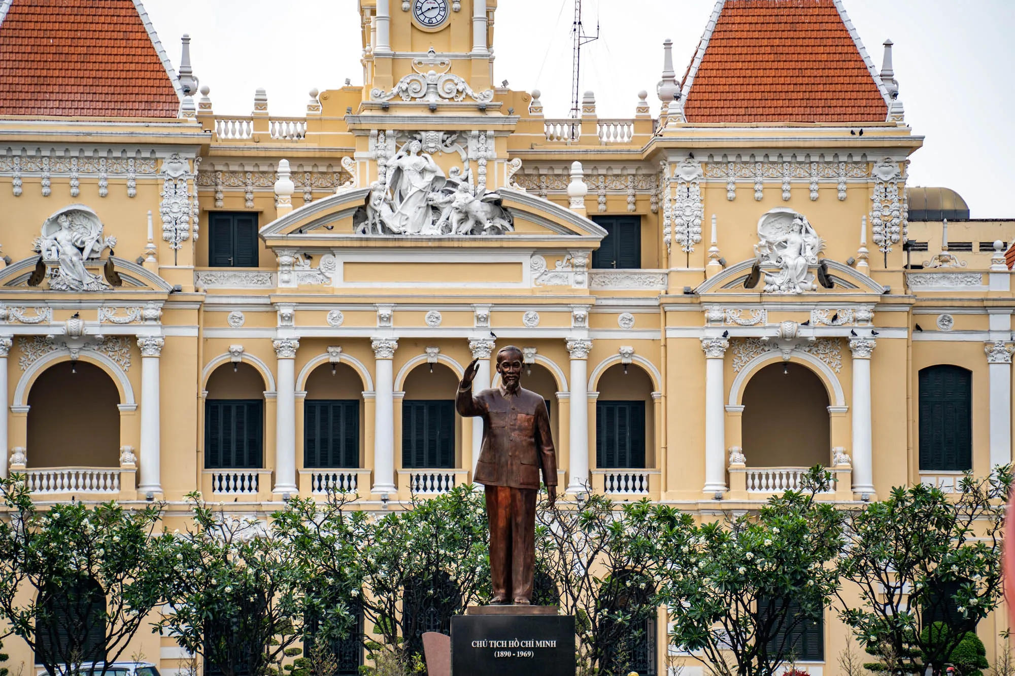この写真はベトナムのホーチミン市で撮影されたものです。画面中央には、ホー・チ・ミンの像が立っています。像は台座の上に立ち、手を上げています。台座には「CHỦ TỊCH HỒ CHÍ MINH (1890 - 1969)」と刻まれています。像の背景には、美しい黄色の建物、ホーチミン市人民委員会庁舎がそびえ立っています。建物はフランス植民地時代の建築様式で、オレンジ色の屋根と白の装飾が特徴的です。建物には、彫刻や時計台が見えます。写真全体は、晴れた日に撮影されたようで、明るい光が当たっています。前景には緑の木々が配置されており、像と建物を引き立てています。全体的に、歴史と文化を感じさせる、印象的な写真です。