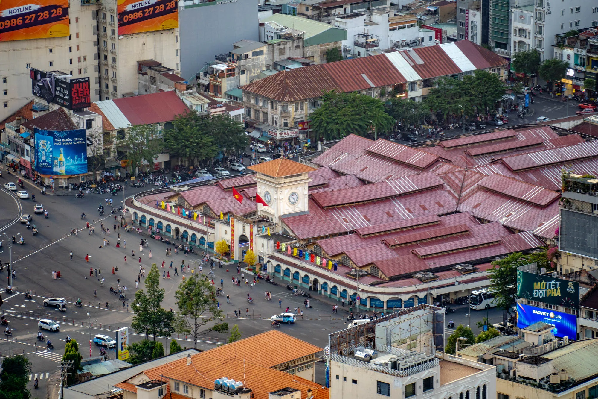 写真はベトナムのホーチミン市を上空から撮影したものです。中心には、特徴的な赤茶色の屋根が連なるベンタイン市場が大きく写っています。市場の前には広場があり、多くの人々が歩いていたり、バイクや車が通行していたりして、活気のある様子が伝わってきます。市場の正面には時計台があり、ベトナムの国旗が掲げられています。  周囲には、様々な高さの建物が立ち並び、広告看板も多く見られます。一部の建物には、屋上庭園のような緑が見え、都市の中に自然を取り入れている様子も伺えます。全体的に、ホーチミン市の賑やかさと活気が感じられる一枚です。