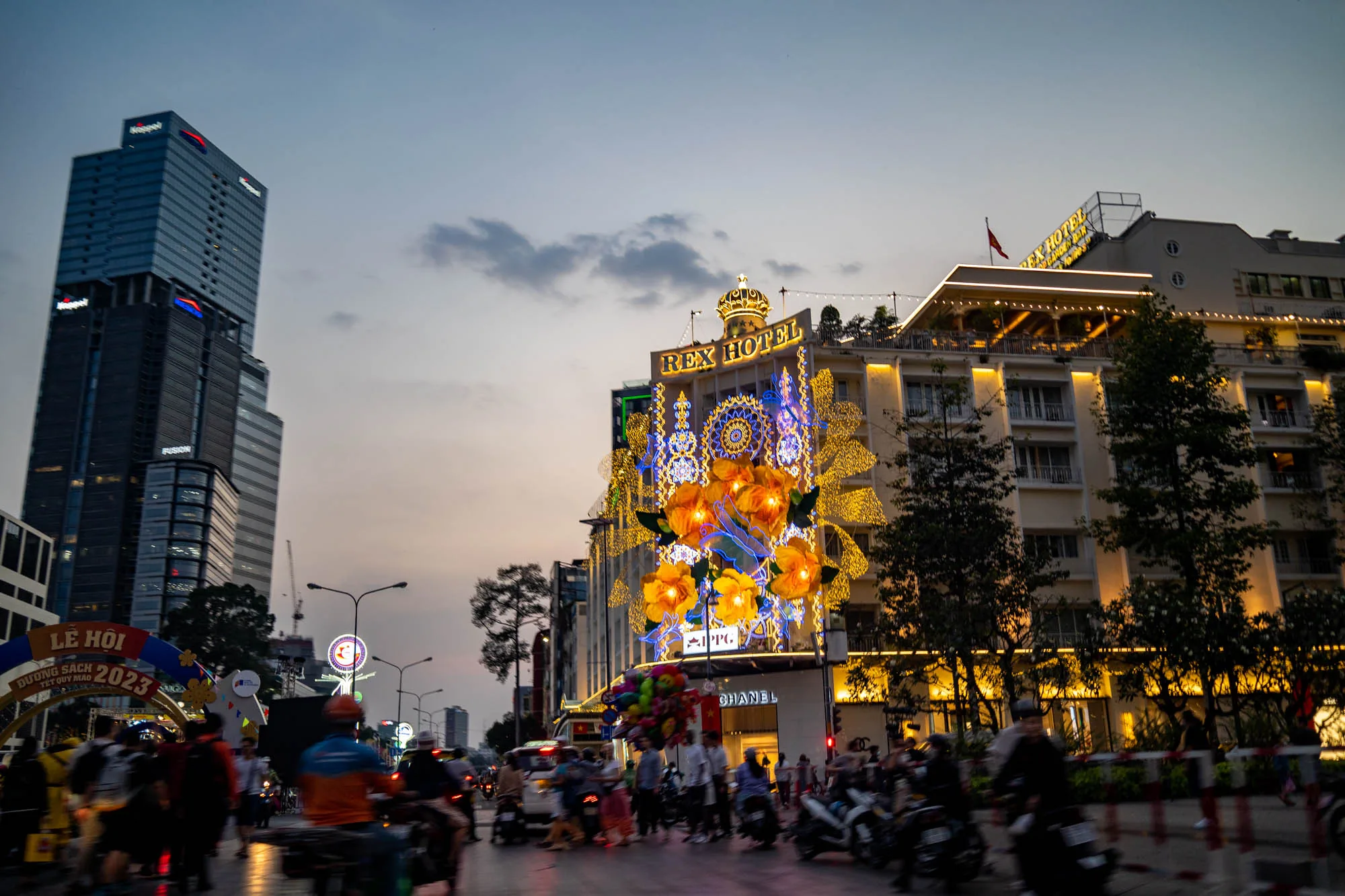 ベトナムのホーチミンで撮影された夜景の写真です。中心にはライトアップされた「REX HOTEL」が大きく写っており、建物全体が華やかな装飾で彩られています。ホテルの正面には、オレンジ色の大きな花のモチーフや、青色の装飾が施され、その下にはCHANELの店舗も見えます。手前には多くの人々やバイクが行き交い、道路の賑わいが感じられます。写真左側には、高いビルがそびえ立っており、現代的な都市の景観も捉えられています。空は夕暮れ時の薄暗い色合いで、全体的に暖色系の光が強調され、活気がありつつも落ち着いた雰囲気を醸し出しています。写真の左端には「Lễ hội Đường Sách Tết Quý Mão 2023」と書かれたアーチがあり、イベントの開催を知らせているようです。