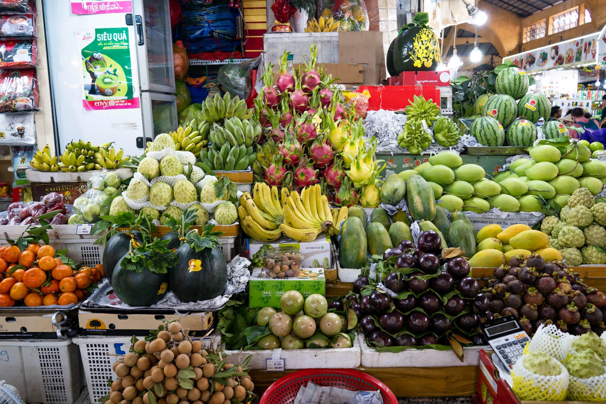 写真はベトナムのホーチミンにある市場の様子を捉えたものです。画面全体に、色とりどりの熱帯の果物が山積みにされています。オレンジ、バナナ、ドラゴンフルーツ、マンゴー、スイカ、マンゴスチン、釈迦頭（シャカトウ）、ロンガンなど、さまざまな種類の果物が豊富に並べられています。果物は種類ごとに丁寧に積み上げられ、その豊かな色彩と整然とした配置が、市場の活気と豊かさを物語っています。  背景には、他の店や買い物客の姿がぼんやりと見え、市場の賑わいを伝えています。電卓が置かれていることから、ここは果物を販売する店のようです。店の上部には蛍光灯が設置されており、店内を明るく照らしています。冷蔵庫にはZespriキウイの広告が貼られていて、商品の多様性を伺わせます。写真全体から、東南アジア特有の活気ある市場の雰囲気が伝わってきます。