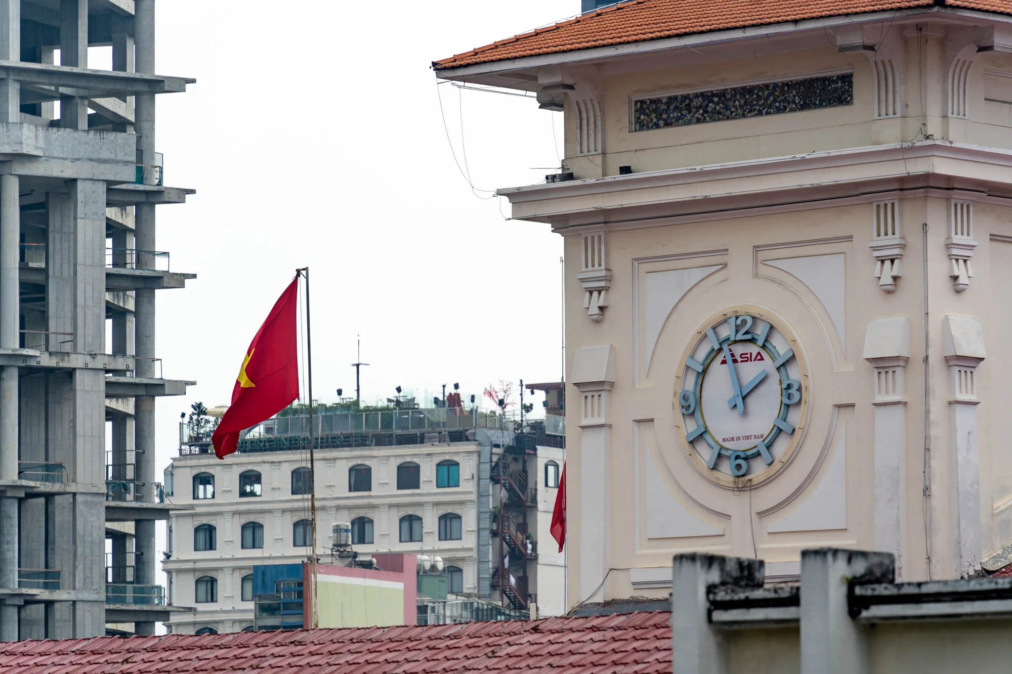 写真はベトナムのホーチミンで撮影されたと思われる風景を捉えています。画面右側には、クリーム色の壁にオレンジ色の屋根を持つ建物の一部が大きく写っており、建物には大きな丸い時計が設置されています。時計の文字盤には「ASIA」の文字と「MADE IN VIET NAM」の文字が確認できます。  画面左側には、建設中のコンクリート製の建物が見え、その手前にはベトナムの国旗が風になびいています。背景には、他の建物もいくつか見えます。  全体的に、ホーチミンの新旧が混在する風景を切り取ったような印象を与えます。曇り空のためか、全体的に落ち着いた雰囲気です。