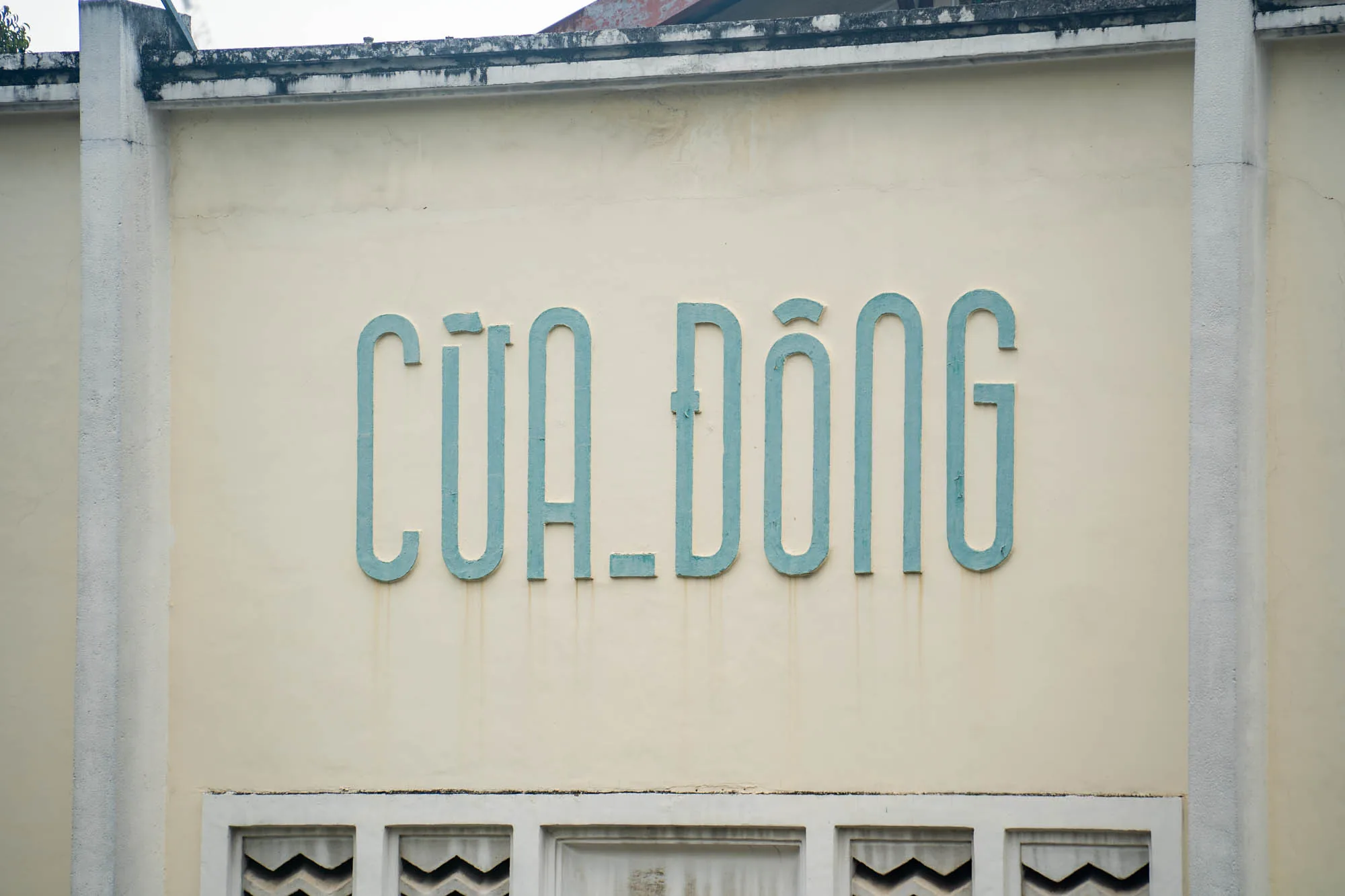 ホーチミンで撮影された、建物の外壁を写した写真です。建物の壁は薄い黄色で、上部には白い枠があります。壁の中央には、水色の太字で「CỬA ĐÔNG」と書かれています。文字の下には、白い装飾的な模様が並んでいます。建物はやや古く、壁には汚れやひび割れが見られます。全体的に落ち着いた雰囲気の写真です。