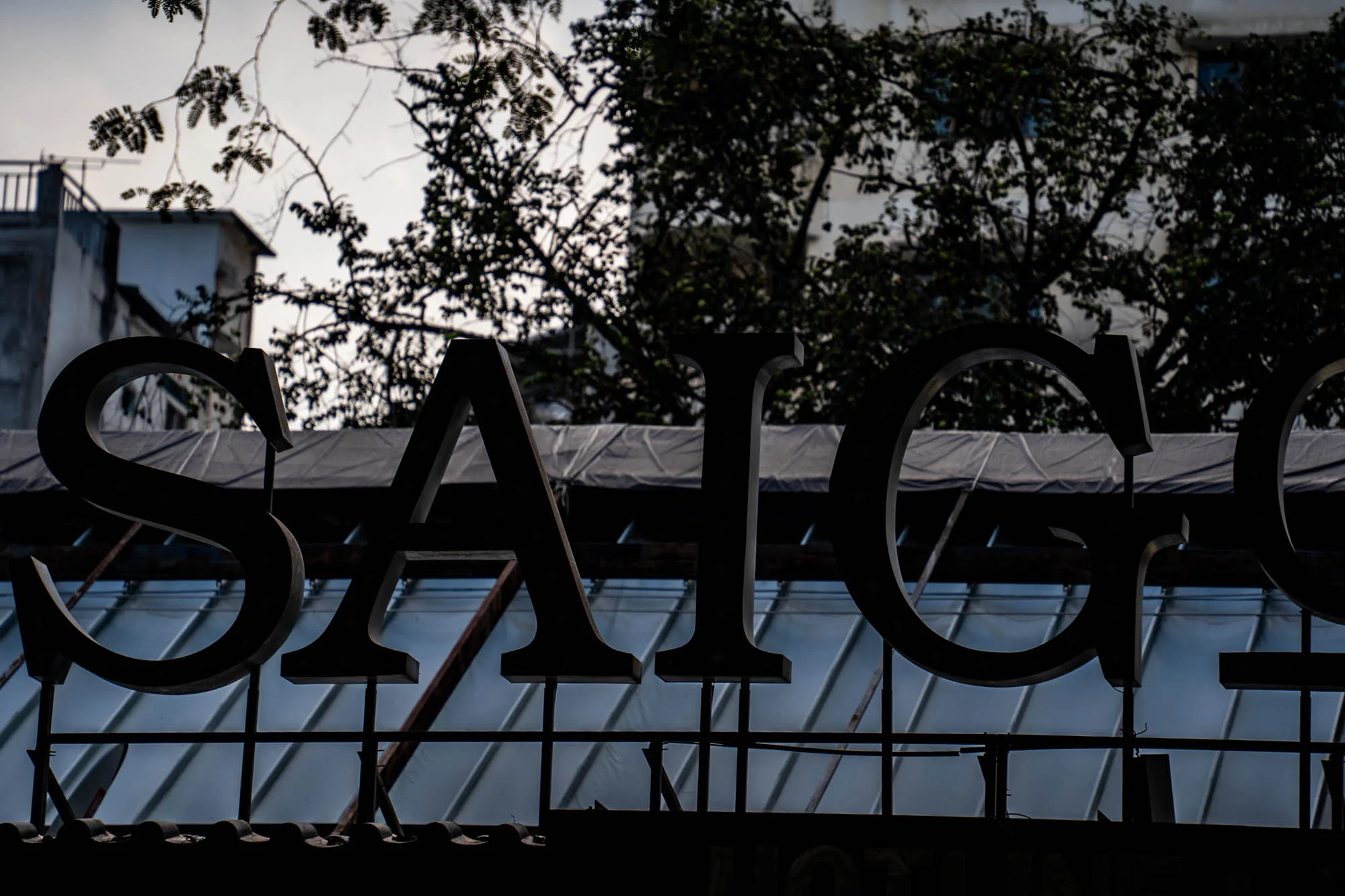 ベトナムのホーチミンで撮影された写真には、「SAIGON」という文字の大きなシルエットが写っています。文字は黒く、背景はやや明るい空です。文字の背後には、建物の輪郭と、葉を落とした木の枝が広がっています。下部にはガラス屋根のようなものが写っており、文字はその上に取り付けられているようです。全体的にモノクロに近い色調で、影が強調され、ノスタルジックな雰囲気を醸し出しています。