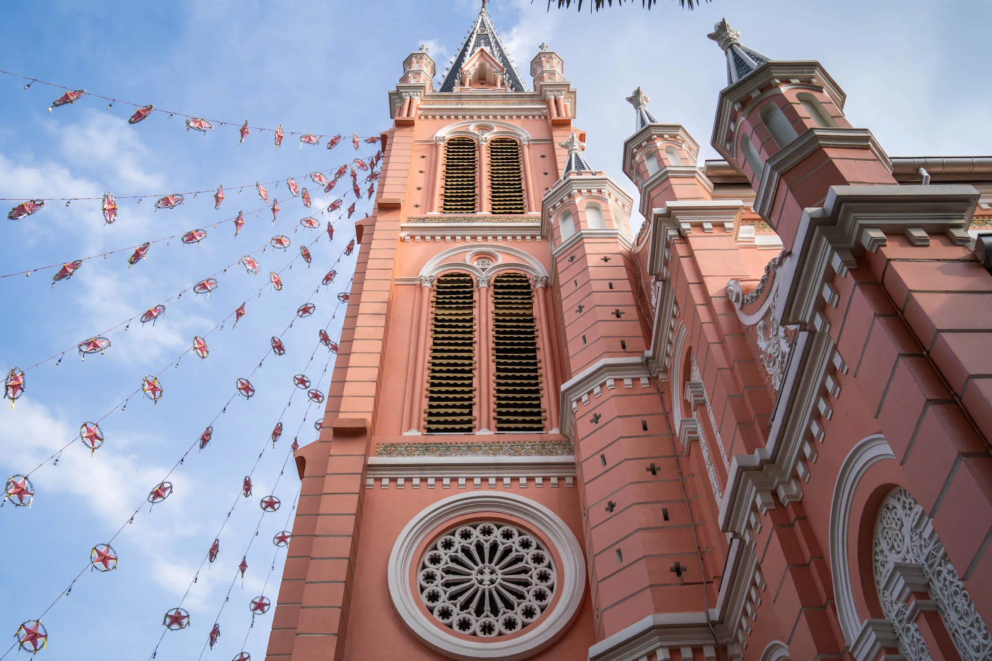 写真はベトナムのホーチミンにある教会の外観を、下から見上げるように撮影したものです。教会全体が特徴的なピンク色で塗られており、ゴシック様式の尖塔や装飾が施されています。建物の側面には小さな十字架の模様が規則的に配置され、美しい装飾が施された円形の窓が見えます。教会の前には、赤い星型の飾りが連なるガーランドが空に張り巡らされており、お祭りや特別な日の雰囲気を醸し出しています。空は薄い水色で、全体的に明るく穏やかな印象です。