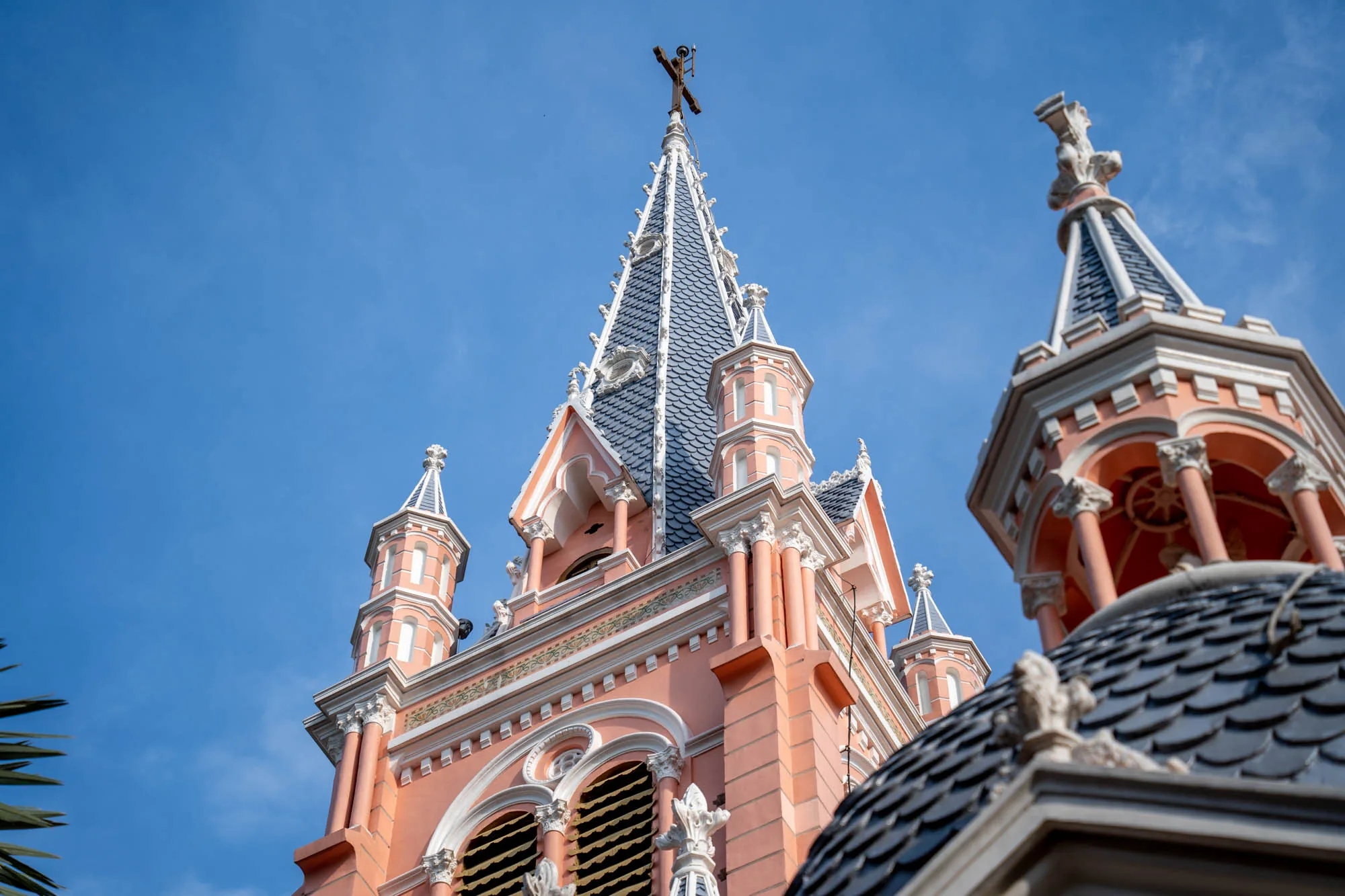 これはベトナムのホーチミンで撮影された写真です。写真には、ピンク色を基調とした美しい教会の建物が写っています。空は晴れており、教会の尖塔が青空に映えています。教会の屋根はダークグレーの瓦で覆われており、その上に十字架が立っています。建物は細部にまで凝った装飾が施されており、建築様式の美しさが際立っています。柱やアーチ、彫刻など、西洋風の要素が取り入れられています。教会の隣にはヤシの葉が見え、南国の雰囲気も感じられます。全体的に明るく、穏やかな印象を与える写真です。