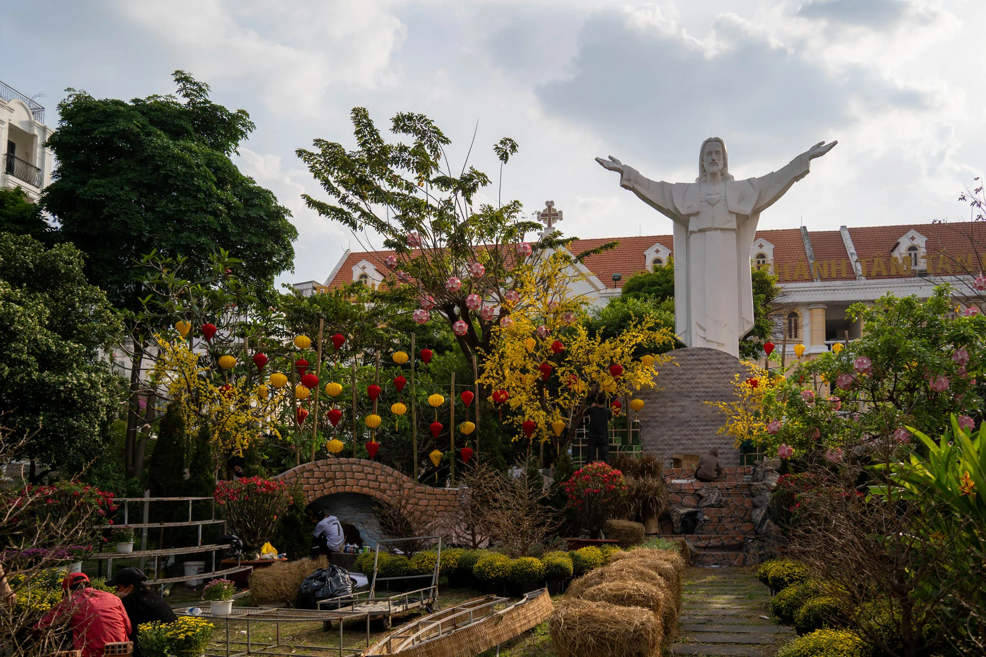 写真はベトナムのホーチミンで撮影されたもので、庭園のような場所を写しています。画面中央やや右には、両手を広げたキリスト像が白く輝いています。その背景には、ヨーロッパ風の建物が見え、赤茶色の屋根が特徴的です。庭園には、黄色や赤色の提灯が飾られた木々が点在しており、ベトナムの旧正月であるテトの飾り付けのようです。  画面左側には、木製の棚や藁で作られたベンチのようなものが置かれており、数人の人が作業をしている様子が見えます。レンガ造りの小さなアーチ橋の下にも人がおり、装飾の準備をしているのかもしれません。全体的に、テトに向けて庭園が美しく飾り付けられている様子が伝わってくる、明るく華やかな雰囲気の写真です。空には雲が広がっており、日差しは柔らかく感じられます。