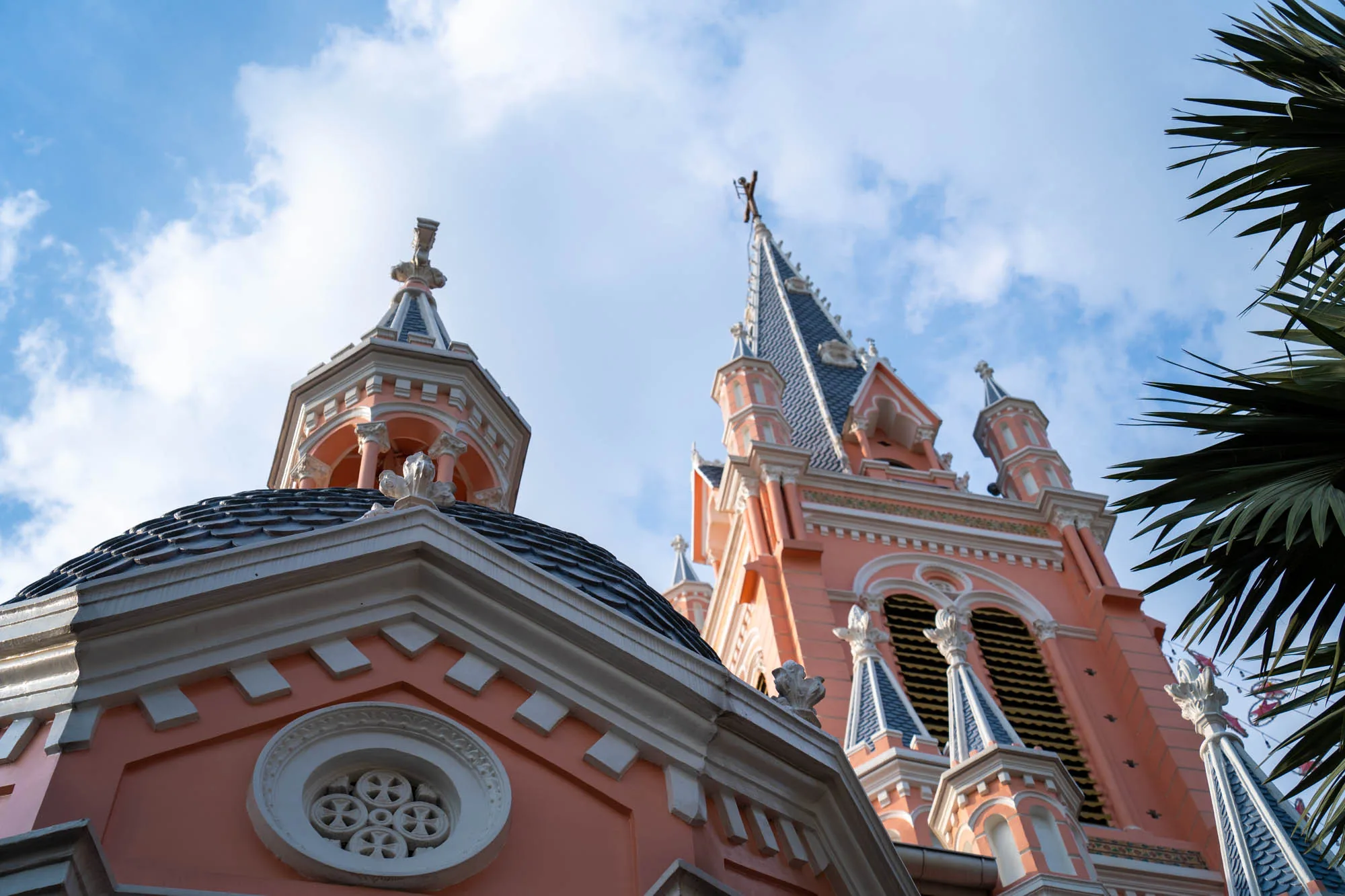 ベトナム、ホーチミンで撮影された写真です。画面いっぱいにピンク色の教会の一部が写っています。建物の外観は複雑な装飾が施されており、淡いピンク色の壁に白い縁取りがされています。丸い屋根の上には小さな塔があり、尖塔の屋根には十字架が立っています。背景には青空が広がり、白い雲が浮かんでいます。写真右端にはヤシの葉が写り込んでおり、トロピカルな雰囲気を添えています。全体的に明るく穏やかな印象を与える写真です。