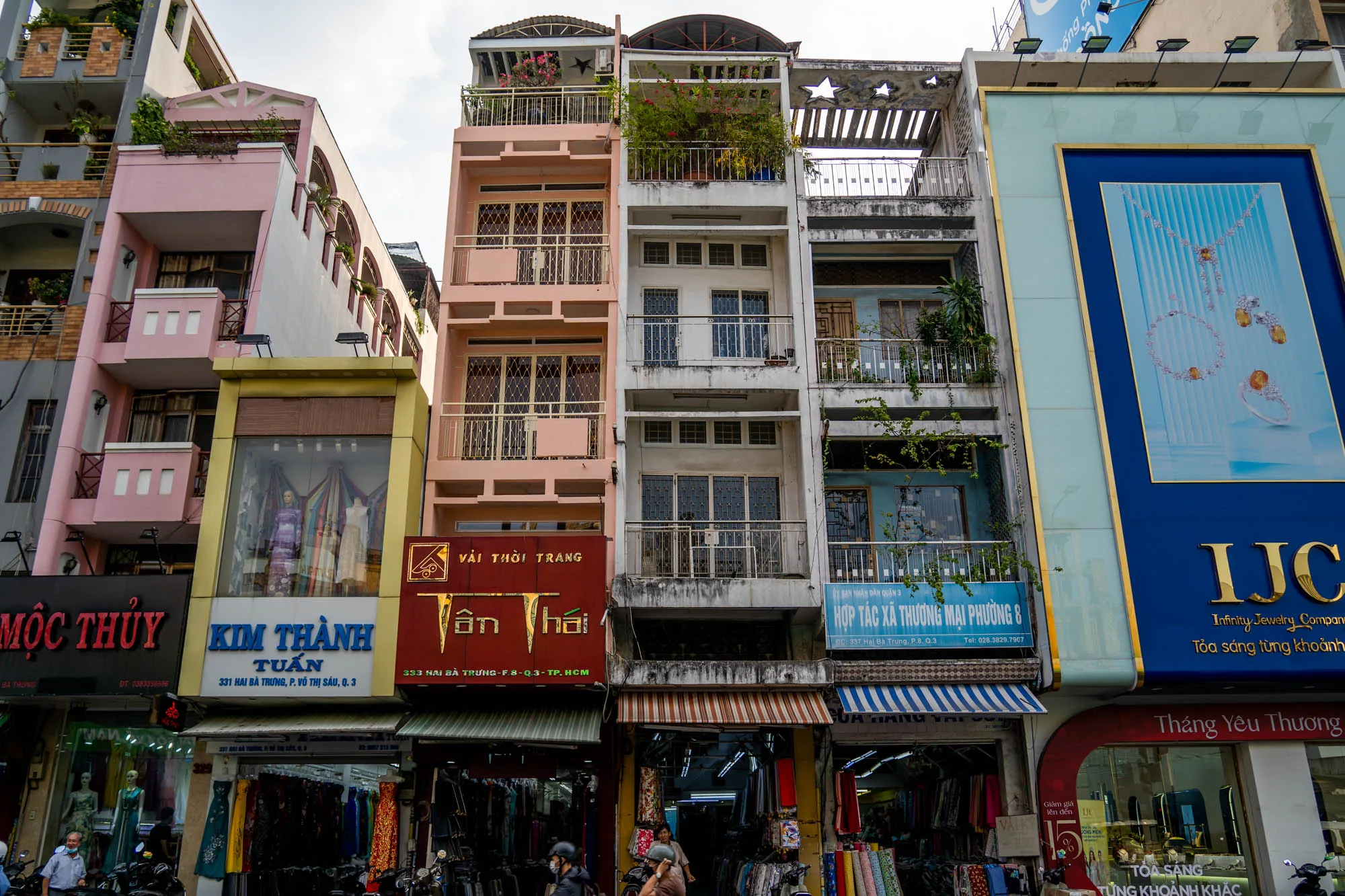 ベトナムのホーチミン市で撮影された写真です。画面には複数の細長い建物が並んでおり、それぞれ異なる色合いで塗られています。これらの建物は、路上に面した店舗とその上の住居が一体となっているようです。一番左の建物はピンク色で、その隣は黄色の看板が目立つ店舗、さらにその隣はピンク色の建物、その隣は白っぽい建物、一番右は水色の看板を掲げた建物となっています。各建物の下部には店舗があり、衣料品やアクセサリーなどを販売しているようです。道路にはバイクが多数駐車されており、人々が行き交う様子も見られます。建物の装飾や看板の文字などから、東南アジア特有の活気ある雰囲気が伝わってきます。