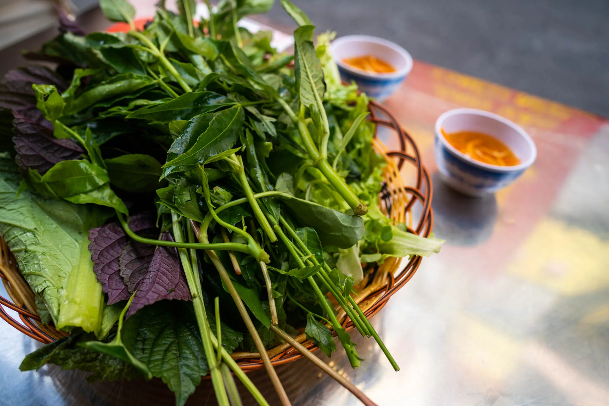 写真はベトナムのホーチミンで撮影された、ベトナム料理の付け合わせと思われる料理のアップです。手前には、様々な種類の緑の葉野菜が盛られた竹製の籠が置かれています。葉野菜は、濃い緑色のものや、紫色のものなど、種類が豊富です。籠の奥には、青い縁取りのある白い器に、オレンジ色のソースが入ったものが二つ並んでいます。テーブルは、赤、黄、白などの色が混ざり合った模様が施されています。全体的に、新鮮な食材の色鮮やかさと、ベトナムの現地の雰囲気が伝わる写真です。