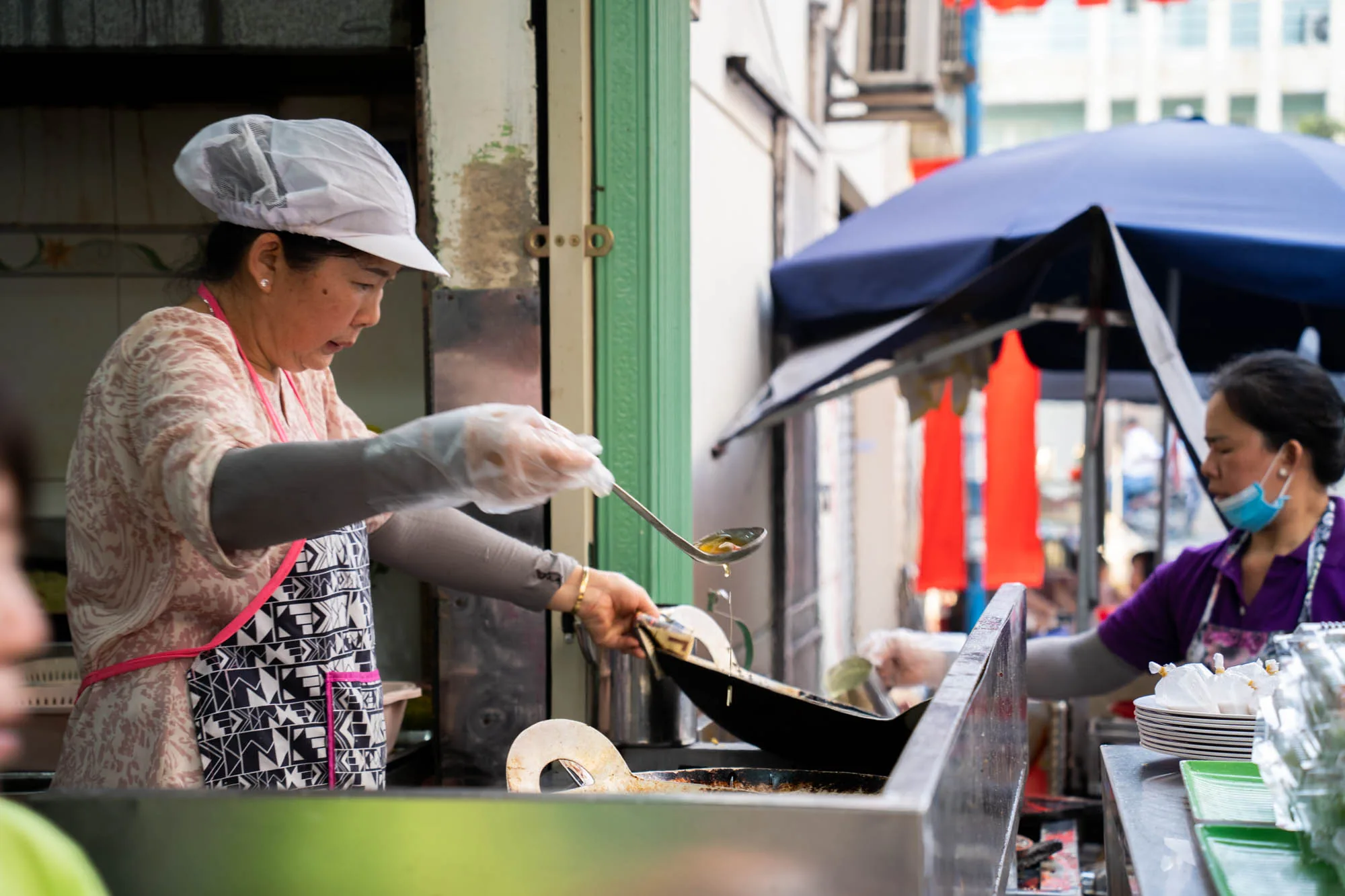 ベトナムのホーチミンで撮影された写真です。路上にある屋台のような場所で、二人の女性が調理をしています。手前にいる女性はコック帽をかぶり、花柄のシャツに幾何学模様のエプロンをしています。彼女は透明な手袋を着用し、お玉で何かをすくい上げて黒い鍋に入れているようです。奥にもう一人女性がいて、紫色のシャツを着てマスクをしています。彼女も何かの作業をしているように見えます。周囲には、屋台の調理器具や食器、赤い装飾などが写っており、活気のあるアジアのストリートフードの様子が伝わってきます。