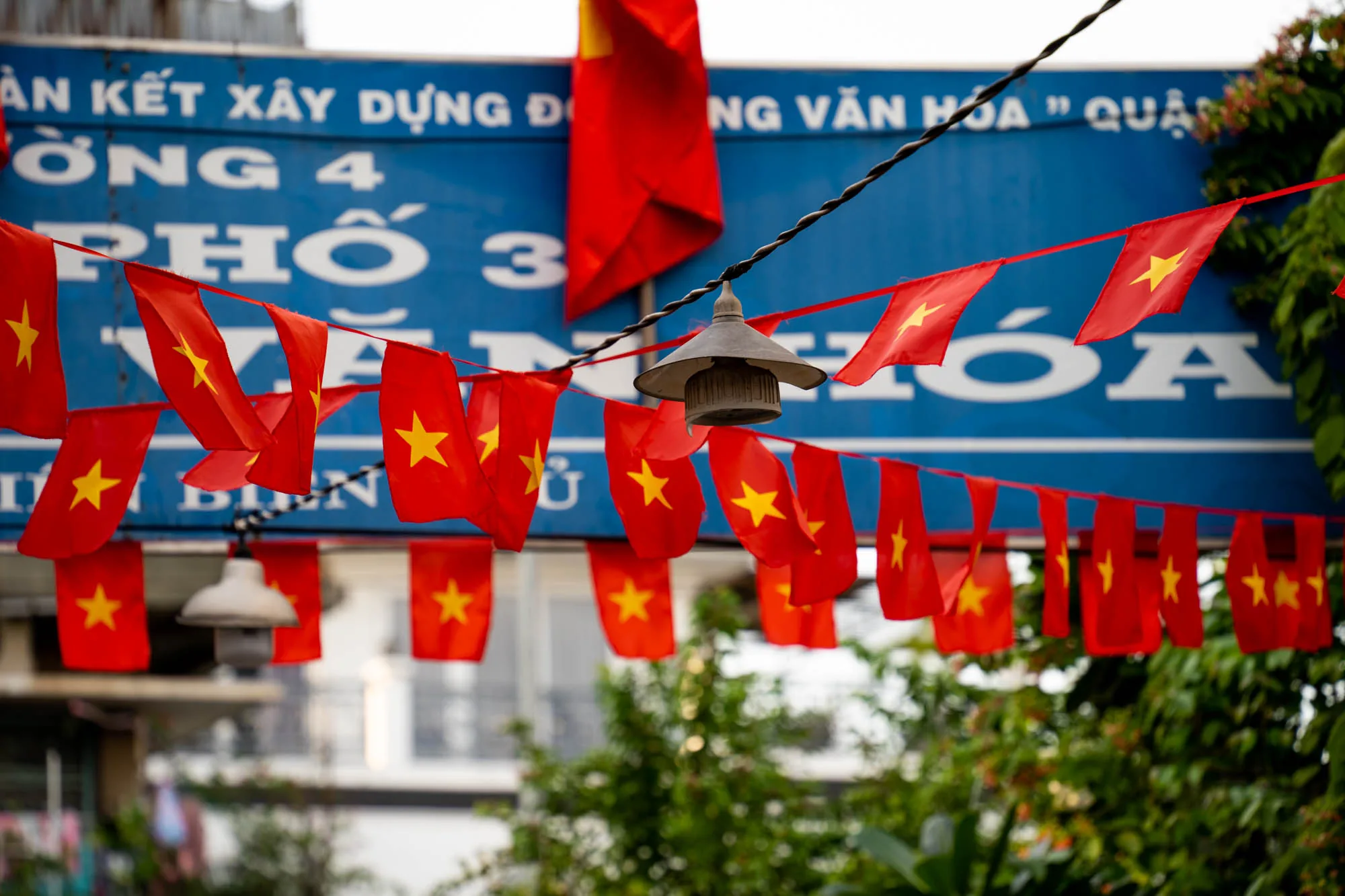 ベトナムのホーチミンで撮影された写真です。画面全体に赤地に黄色の星が描かれたベトナムの国旗のミニチュア版が、紐に連なって飾られています。国旗は道路を横断するように張られており、風になびいています。旗の背景には、青地に白い文字でベトナム語の看板が見えます。看板には「ĐOÀN KẾT XÂY DỰNG ĐỜNG 4 PHỐ 3 NG VĂN HÓA " QUẬ」と書かれています。電線や街灯も写っており、生活感のある街の風景を切り取っています。画面下部には緑の木々が写り込み、自然の要素も感じられます。写真全体はやや霞んでおり、柔らかい光が差しているように見えます。街の活気とベトナムの愛国心が感じられる写真です。