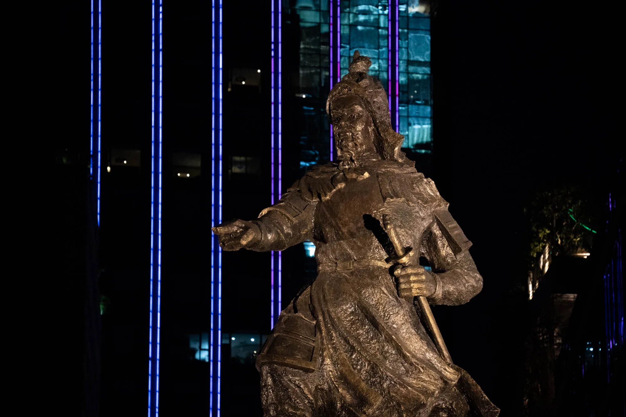 ホーチミン市で撮影された写真です。夜間の撮影で、暗い背景の中に英雄の像が浮かび上がっています。像は、鎧を身に着け、剣を持ち、指を指し示す姿で、威厳と力強さを感じさせます。像の素材はブロンズのようであり、表面の質感や細部が丁寧に表現されています。背景には、青と紫の光でライトアップされた高層ビルがそびえ立ち、都会的な雰囲気と、歴史的な像の対比が印象的です。周囲は暗く、像とビルの光が際立っています。全体として、ホーチミン市の歴史と現代性が融合した様子が捉えられた写真です。