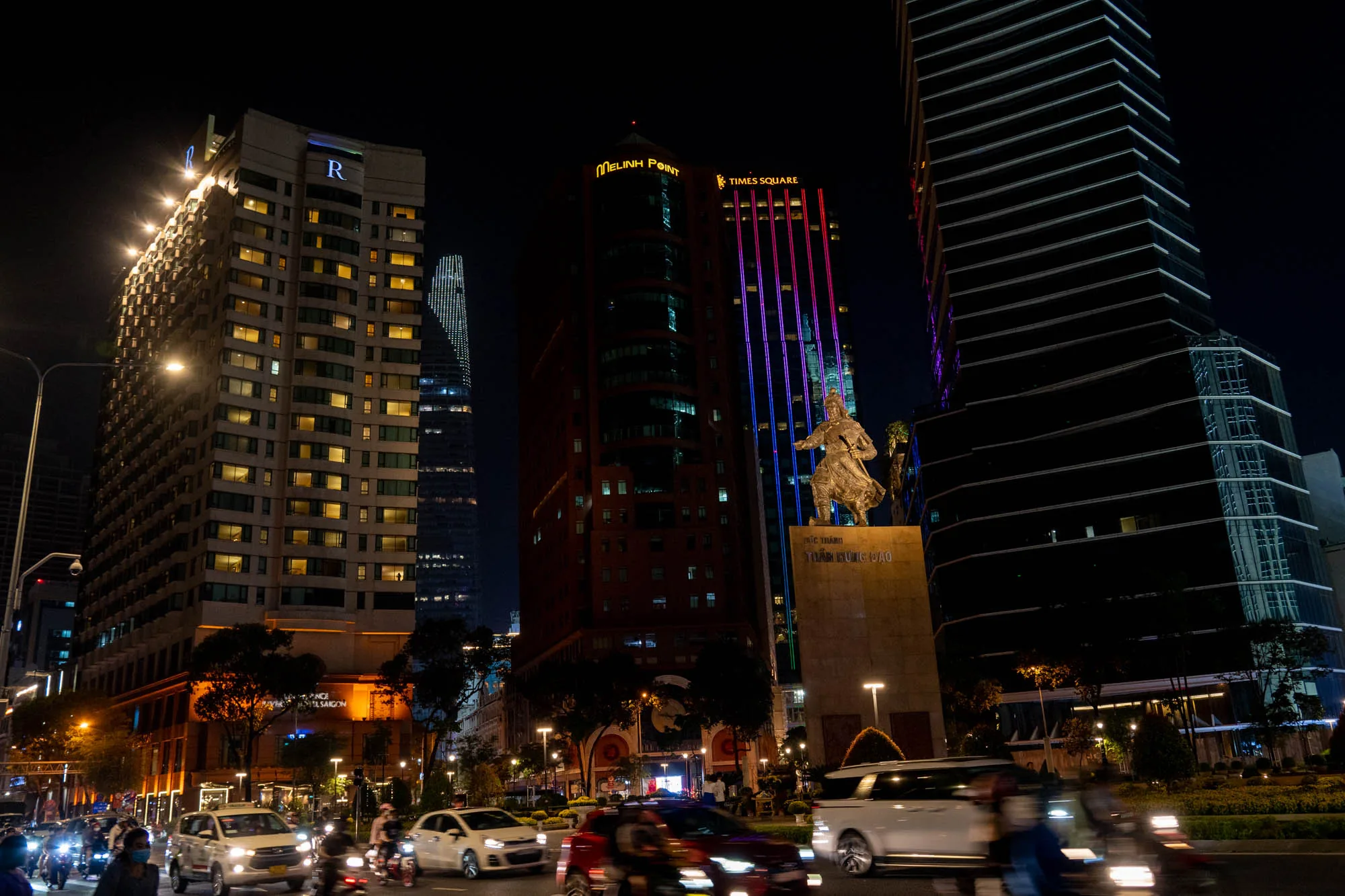 この写真はベトナムのホーチミン市で撮影された夜景です。画面中央にはチャン・フン・ダオ将軍の像がライトアップされており、その背後には高層ビル群がそびえ立っています。左側には「R」のロゴが光るホテルがあり、多数の窓から光が漏れています。中央には「Melinh Point」と「Times Square」の文字が輝くビルがあり、ピンクと青のライトが垂直に走っています。右側には黒くモダンなデザインのビルがそびえています。  手前の道路には車やバイクがひっきりなしに行き交い、都会の賑やかさを感じさせます。車のライトや街灯が光り、夜の街を照らしています。写真全体は暗い色調ですが、それぞれの建物のライトアップが鮮やかで、コントラストが効いた美しい夜景を作り出しています。