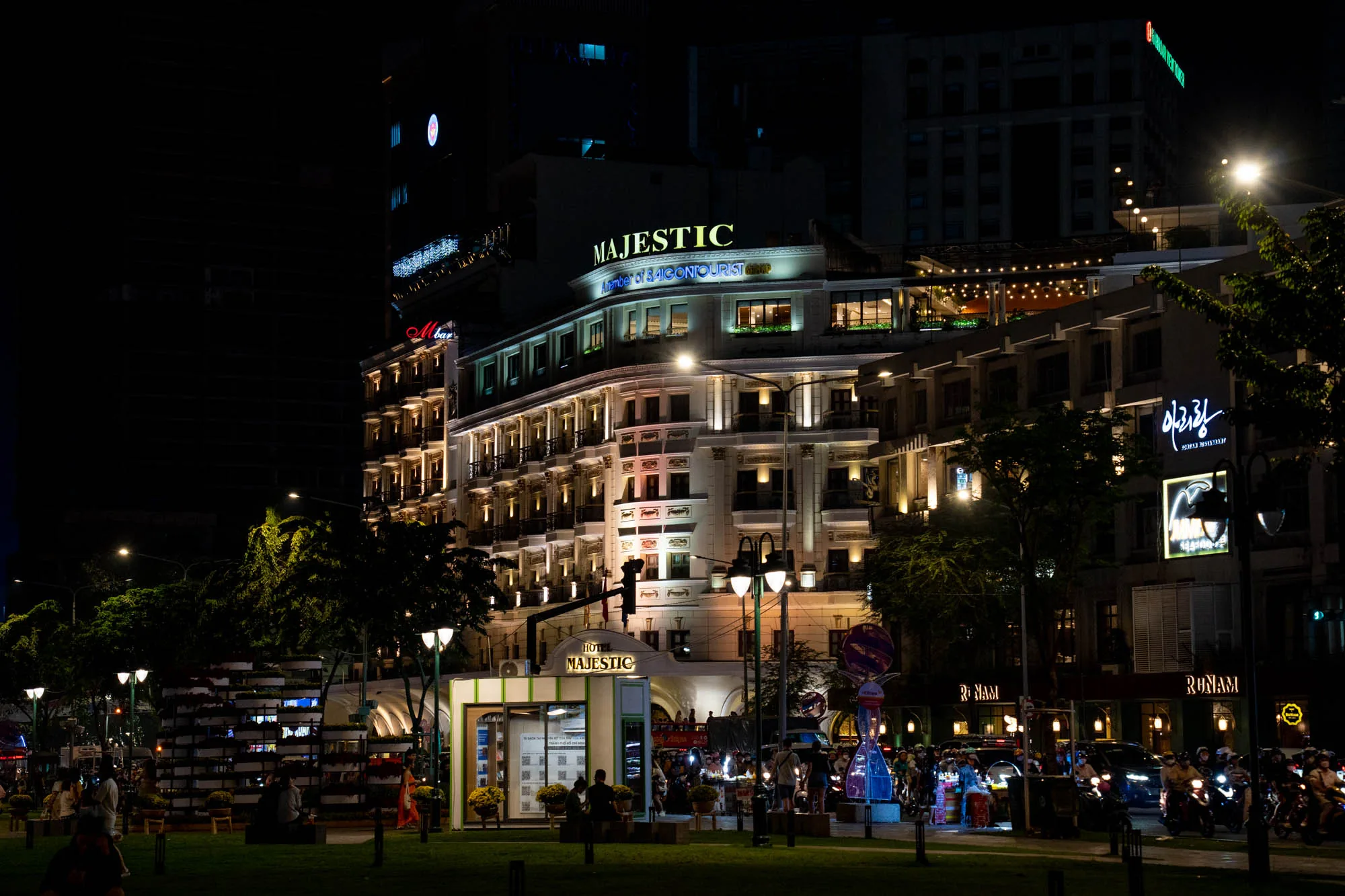 ベトナムのホーチミン市で撮影された夜景の写真です。中央にはライトアップされた「MAJESTIC」と書かれたホテルがあります。ホテルの外観は白く、バルコニーと窓枠がライトで縁取られ、クラシカルで重厚な雰囲気を醸し出しています。ホテルの手前には芝生の広場があり、街灯が点在しています。広場には人々が座って談笑したり、歩いたりしている様子が見られます。道路には多くのバイクが走っており、街の活気が伝わってきます。背景には高層ビルがそびえ立ち、夜空に浮かび上がっています。写真全体は暖色系のライトで照らされ、都会の夜の賑やかさと落ち着いた雰囲気が調和しています。右側には韓国料理店の看板がライトアップされています。