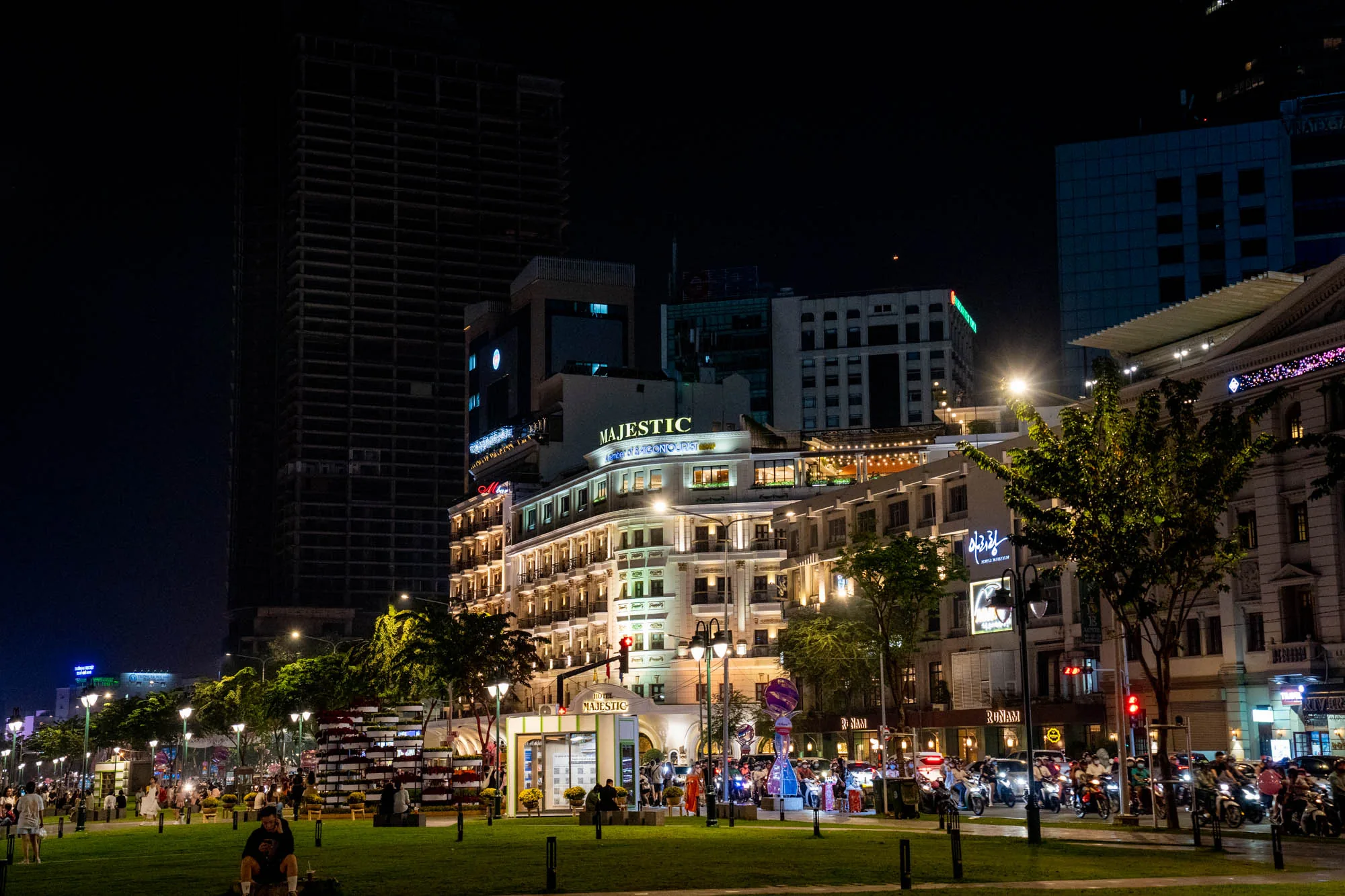 写真はベトナムのホーチミンで撮影された夜景です。画面中央には「Majestic」と書かれたホテルが輝き、その周辺には複数の高層ビルがそびえ立っています。左奥のビルは未完成で、骨組みだけが見えています。  写真手前には緑豊かな公園があり、人々がベンチに座ったり、歩いたりして過ごしています。道には多くのバイクが停車しており、ホーチミンの活気ある夜の雰囲気を伝えています。街灯が並び、温かい光を放ち、夜の公園を照らしています。  全体的に、写真はホーチミンの賑やかな夜の様子を捉えており、高層ビルと緑豊かな公園が共存する様子が印象的です。