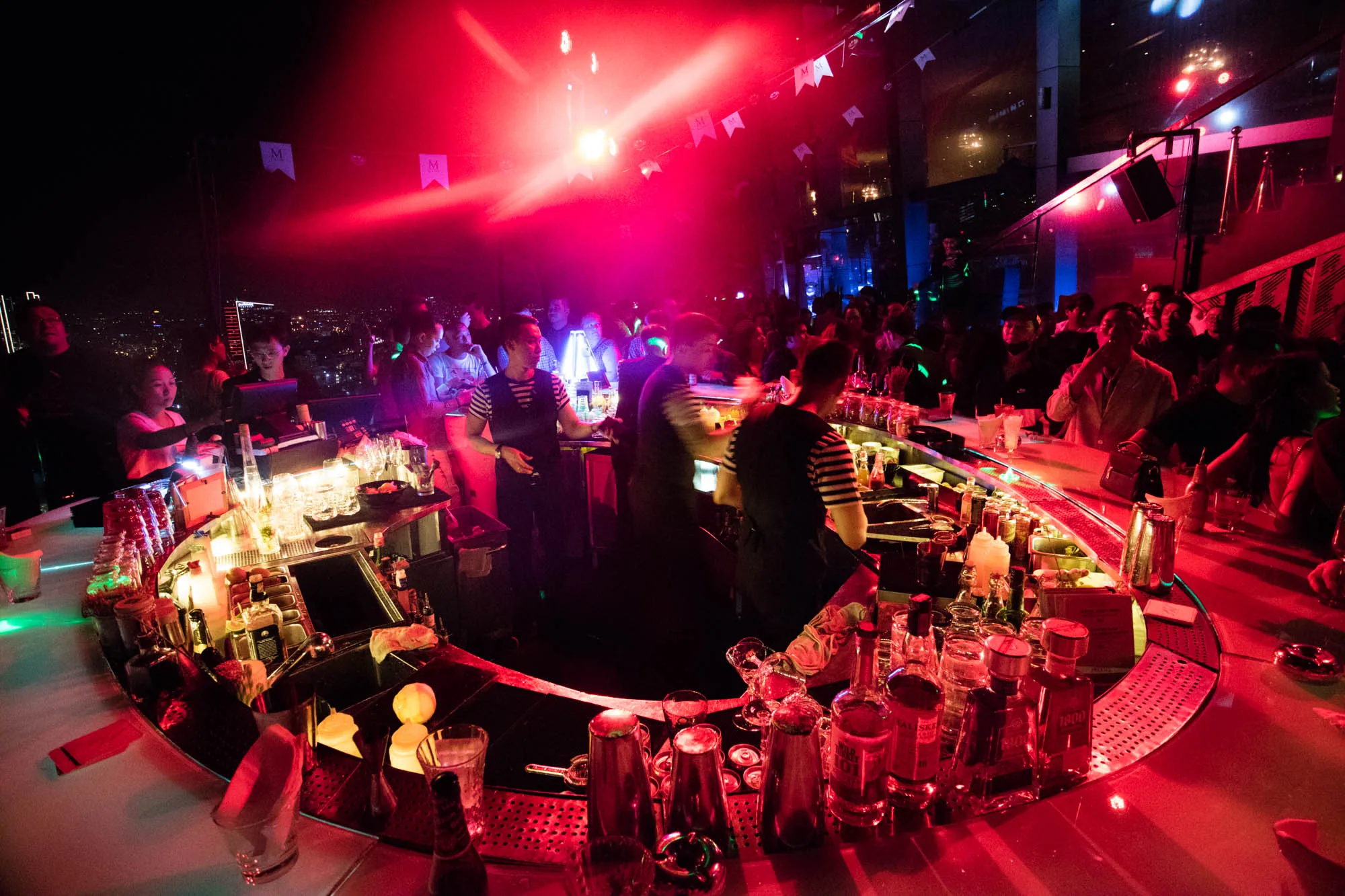 ベトナムのホーチミンで撮影されたと思われる写真です。夜景を背景にした高層階のバーの様子が捉えられています。赤い照明が全体を照らし、賑やかで活気のある雰囲気を作り出しています。バーカウンターは円形で、多数の酒瓶やグラスが並べられています。バーテンダーはストライプのシャツを着ており、忙しそうに働いています。多くの人々がバーカウンターを囲み、談笑したり、飲み物を楽しんだりしています。窓の外にはホーチミンの夜景が広がり、都会の煌びやかさを感じさせます。天井には「M」のロゴが入った旗が飾られており、装飾にもこだわっていることが伺えます。全体的に、お洒落で洗練されたバーの雰囲気が伝わってきます。