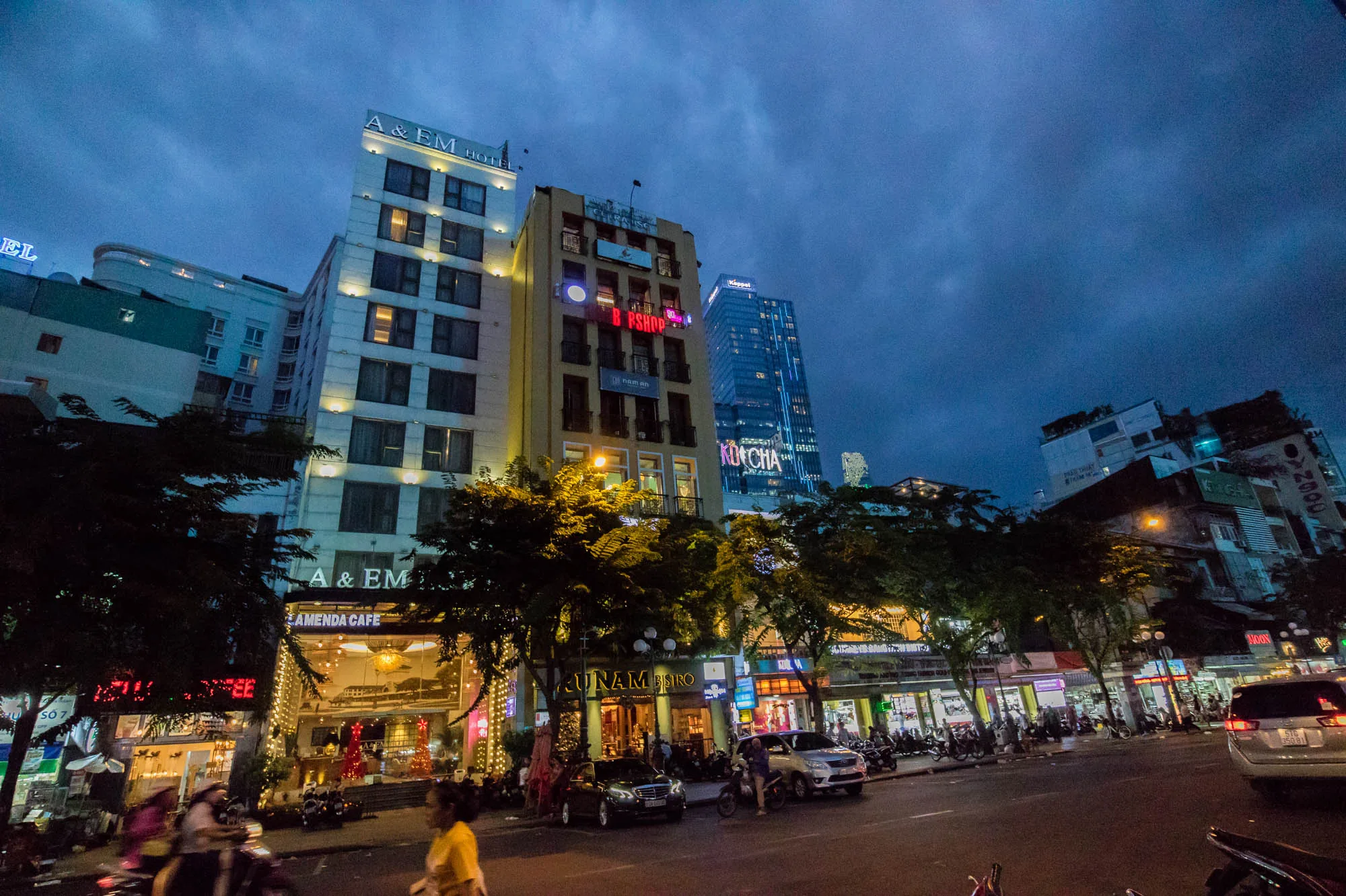 ベトナムのホーチミンで撮影された夜景の写真です。空は暗い青色で、雲が広がっています。写真には、複数の建物が写っており、それぞれ異なる高さとデザインを持っています。左側には「A&EM Hotel」という白い建物が見え、その下には「AMENDA CAFE」の看板があります。中央には「B PSHOP」という看板のある黄色い建物があります。その後ろには、高くそびえ立つガラス張りのビル「KOICHA」がライトアップされています。道路には車やバイクが走行しており、多くの人が行き交っています。街路樹が立ち並び、店舗の照明やネオンサインが夜の街を照らしています。賑やかで活気のある雰囲気です。