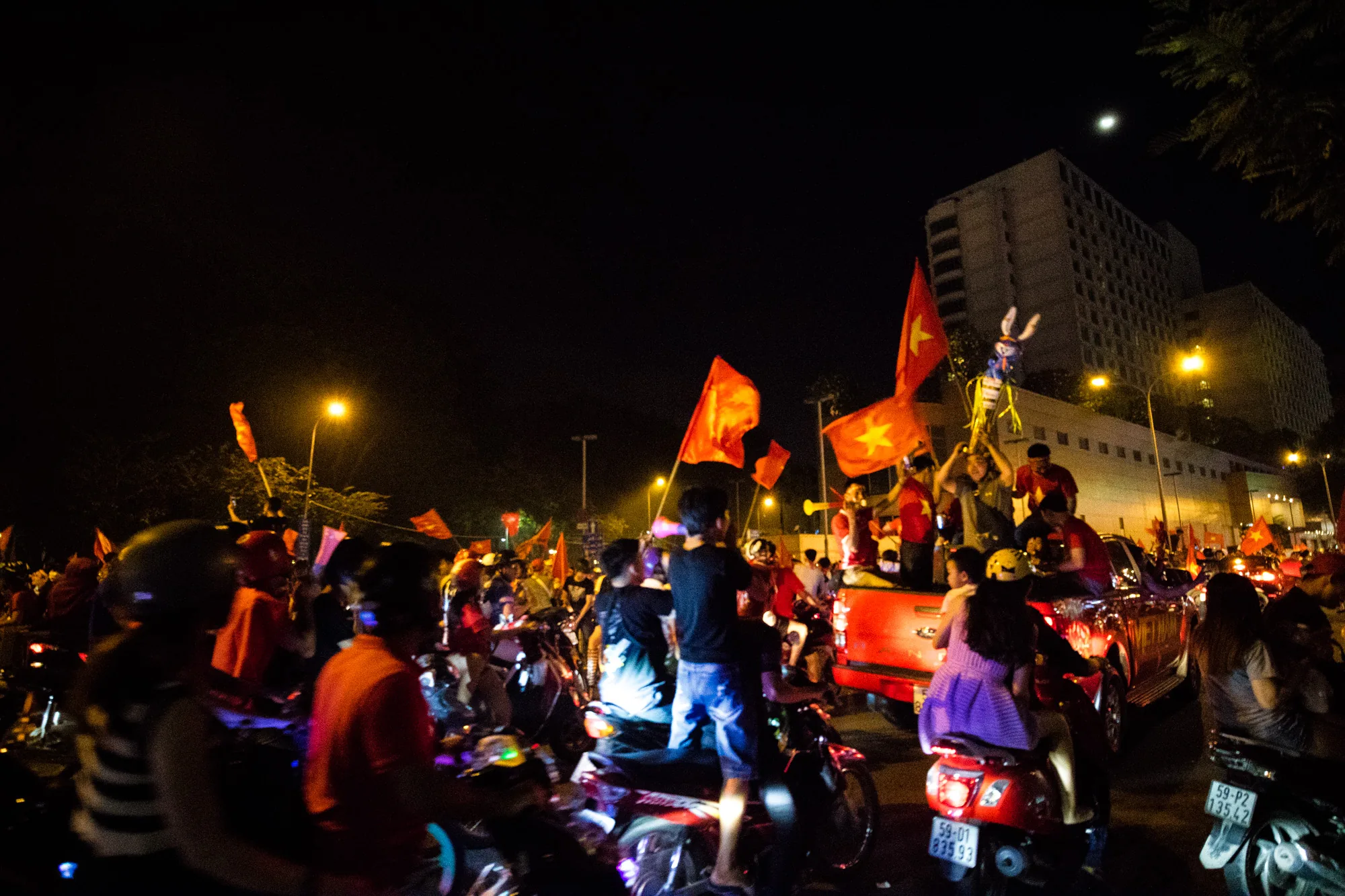 写真はベトナムのホーチミン市で撮影された夜の様子を捉えています。夜空の下、大勢の人々がバイクに乗り、ベトナム国旗を掲げて歓喜に沸いています。街灯の光が人々やバイクを照らし出し、お祭り騒ぎのような熱狂的な雰囲気を醸し出しています。ピックアップトラックの荷台に乗り込み、旗を振ったり、何かを掲げたりして喜びを分かち合っている人々の姿も見られます。背景には高層ビルがそびえ立ち、都会の夜景を彩っています。全体的に、勝利を祝う人々の興奮と高揚感が伝わってくる写真です。