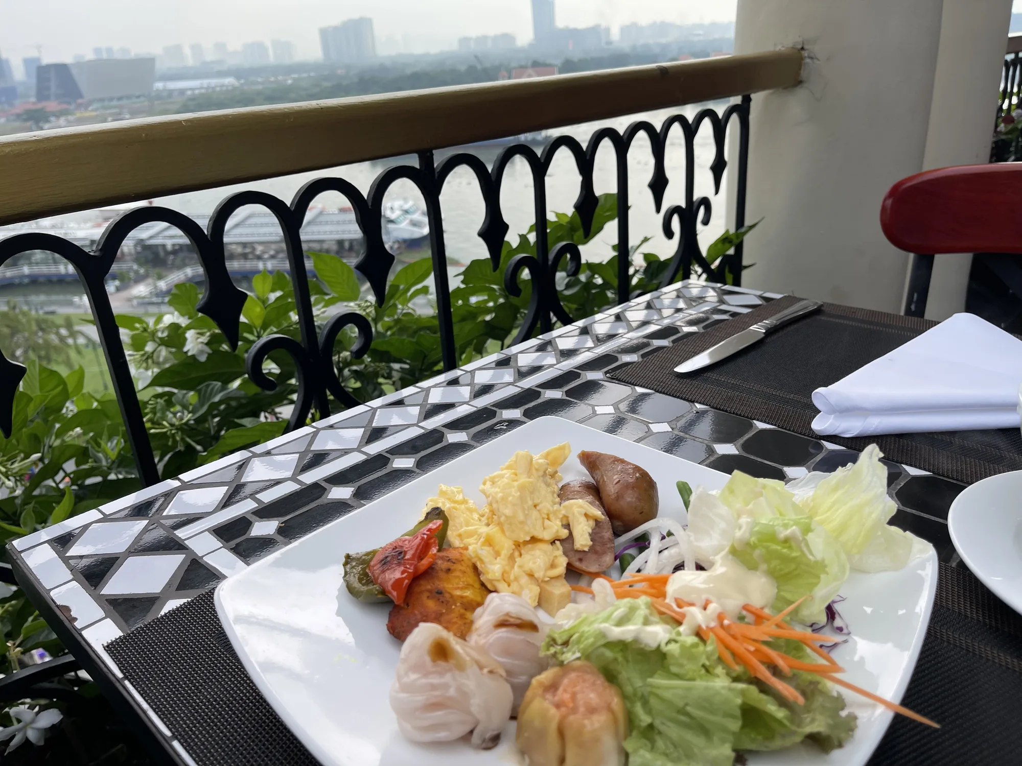 ベトナムのホーチミンで撮影されたと思われる写真です。バルコニーのような場所で朝食を摂っている様子が写っています。画面手前には、白と黒のモザイク模様のテーブルがあり、その上に黒いランチョンマットが敷かれています。マットの上にはナイフとフォーク、白いナプキンが置かれています。テーブルの中央には白い皿があり、スクランブルエッグ、ソーセージ、焼き野菜、シュウマイ、サラダなどが盛り付けられています。バルコニーの柵は黒い鉄製で、植物が茂っており、遠くにはホーチミンの街並みが見えます。曇り空で少し霞がかっており、落ち着いた雰囲気です。