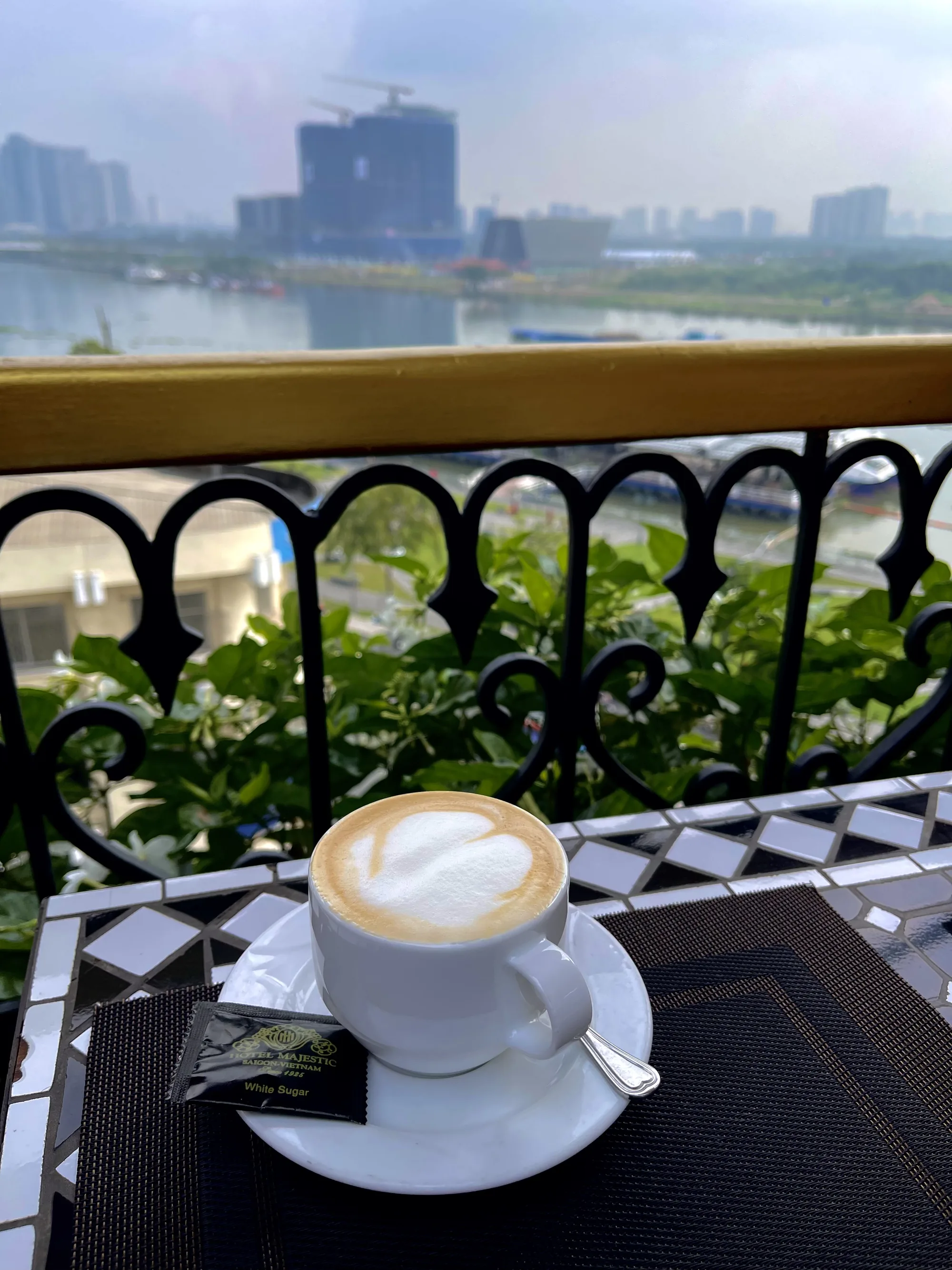 写真は、ベトナムのホーチミン市にあるカフェで撮影されたものです。前景には、白いカップに入ったラテが置かれています。ラテの上には、ハート形の美しいラテアートが描かれています。カップは白いソーサーに置かれ、ソーサーには砂糖の小袋と小さなスプーンが添えられています。カップの手前には、黒いランチョンマットが敷かれています。テーブルは白と黒のモザイクタイルで装飾されています。背景には、植物が植えられたバルコニーがあり、その向こうには川と都市のスカイラインが広がっています。空は曇っており、全体的に穏やかで落ち着いた雰囲気が漂っています。