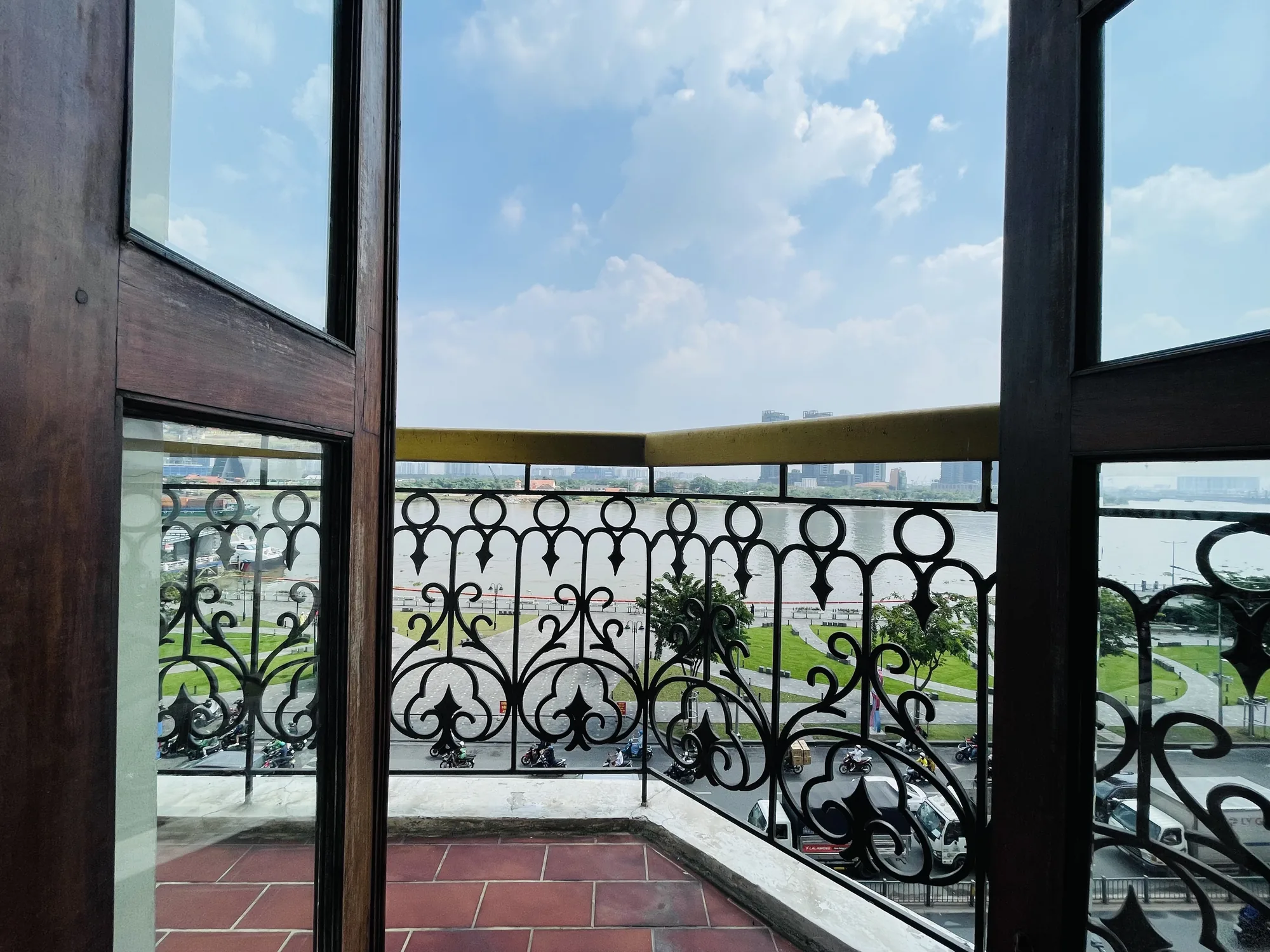 写真はベトナムのホーチミンで撮影されたもので、建物の中から外の景色を眺めている様子を捉えています。画面手前には、レンガ敷きの床と装飾的な鉄格子のバルコニーが見えます。鉄格子の向こうには、川が広がっており、対岸には建物が立ち並ぶ街並みが見えます。空は晴れており、白い雲が浮かんでいます。通りにはバイクや車が通行しており、ホーチミンの活気が感じられます。全体的に、美しい景色と都会の喧騒が組み合わさった、落ち着いた雰囲気の写真です。